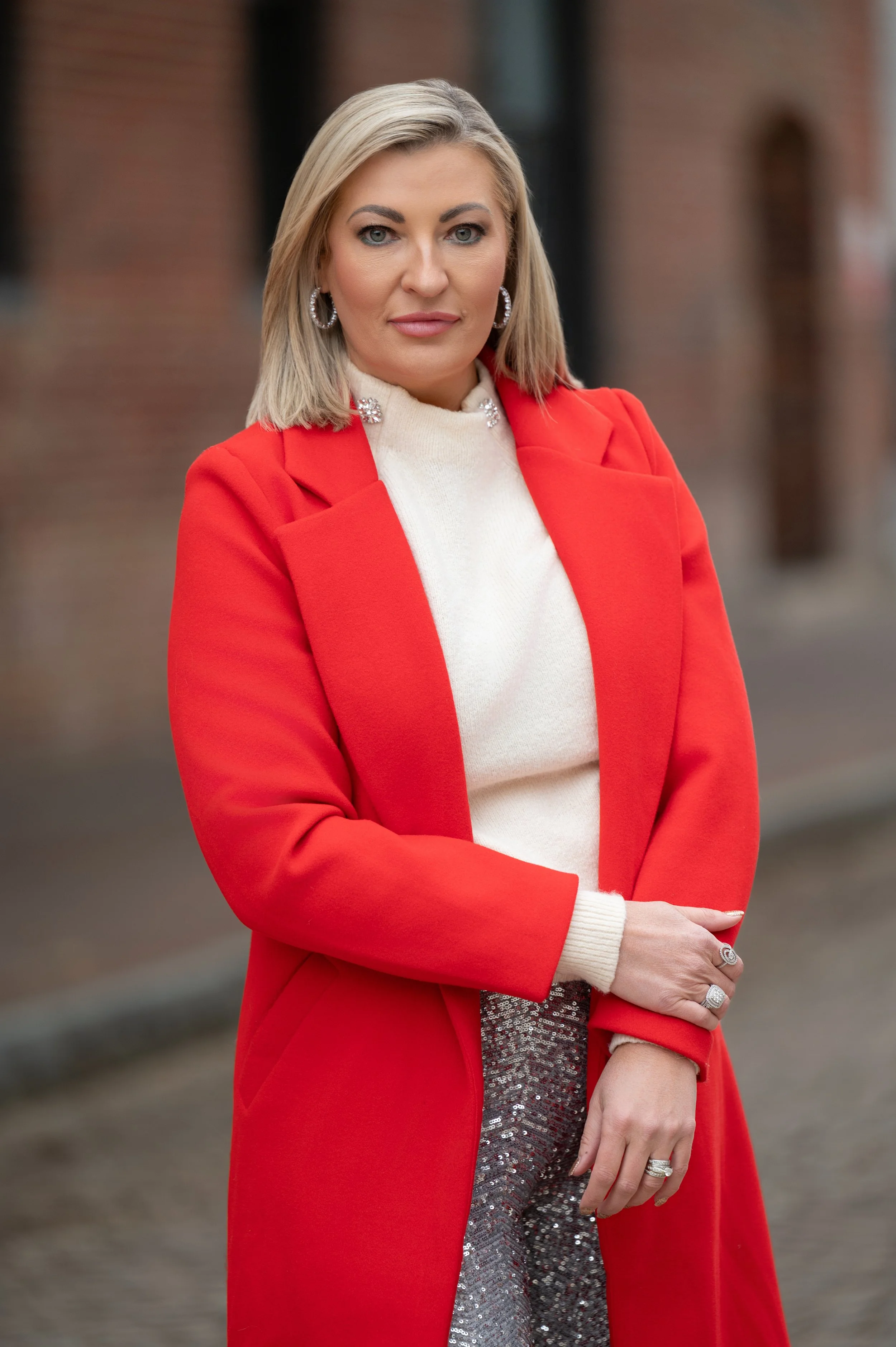 A woman with blonde hair wearing a red coat over a white turtleneck, standing outdoors with a brick building in the background.
