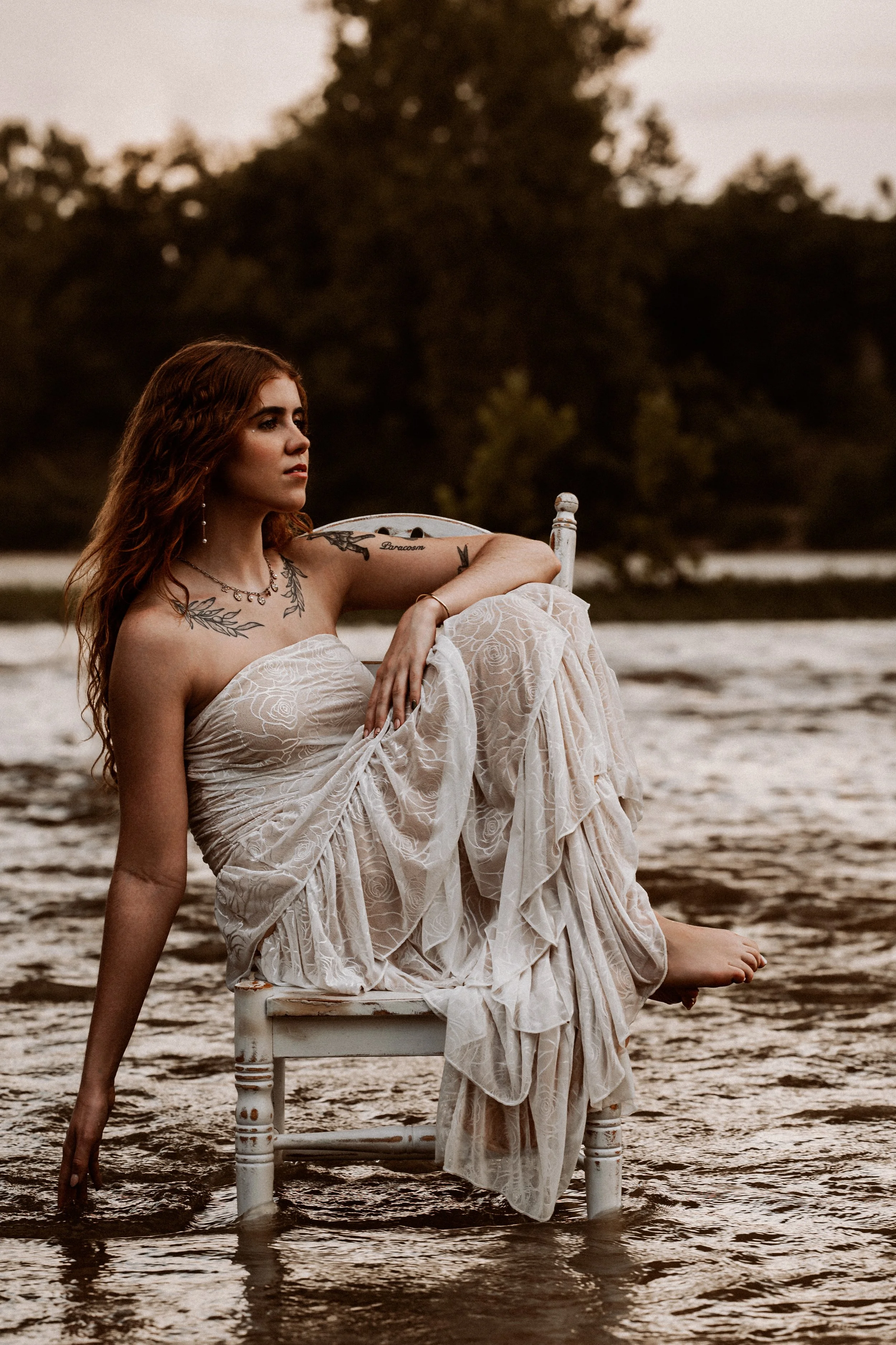 A woman with long red hair wearing a strapless white dress sitting on a white wooden chair partially submerged in water during sunset, with trees in the background.