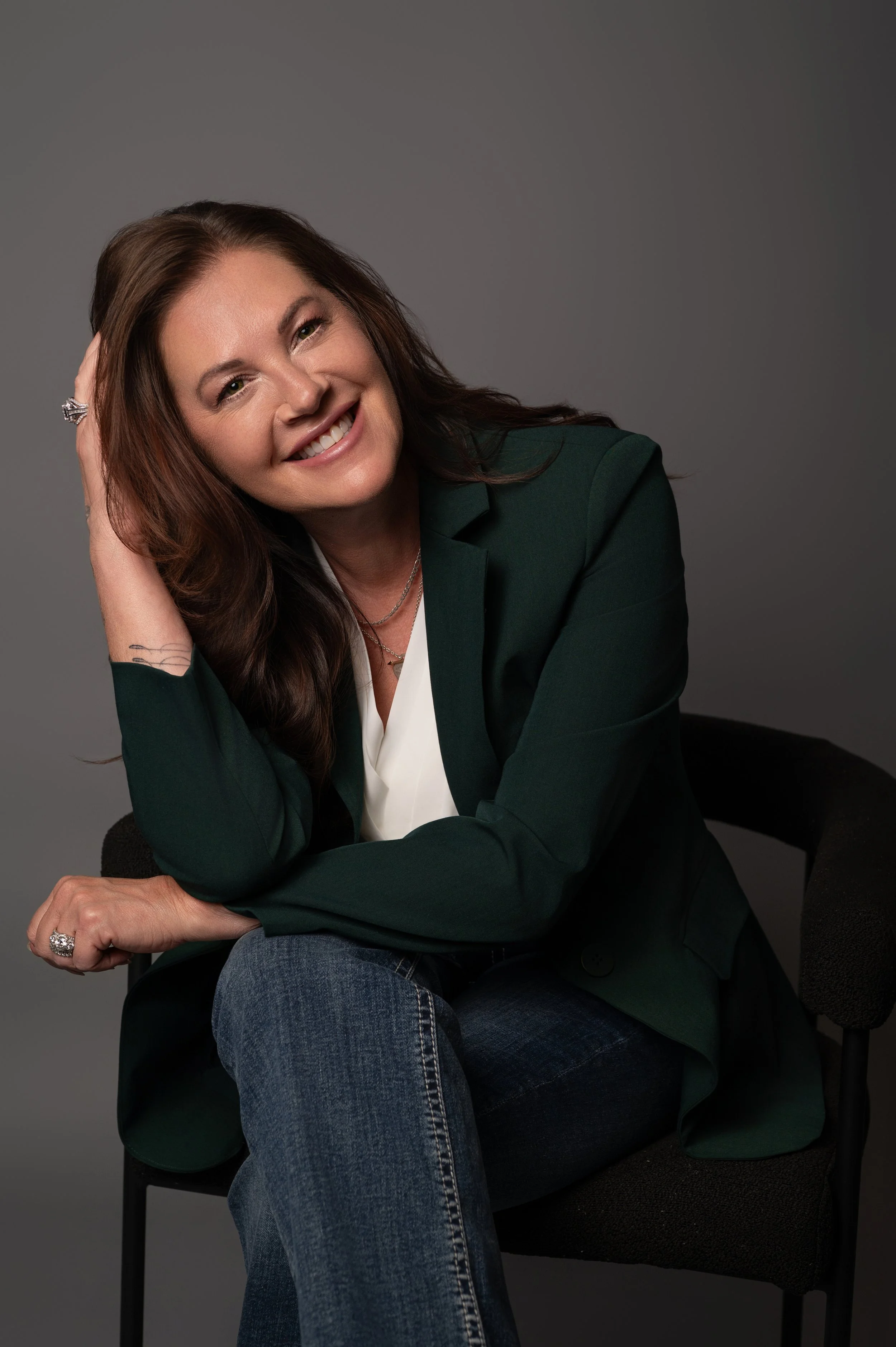 A woman with long brown hair smiling, wearing a dark green blazer, white top, and jeans, seated on a black chair against a gray background.