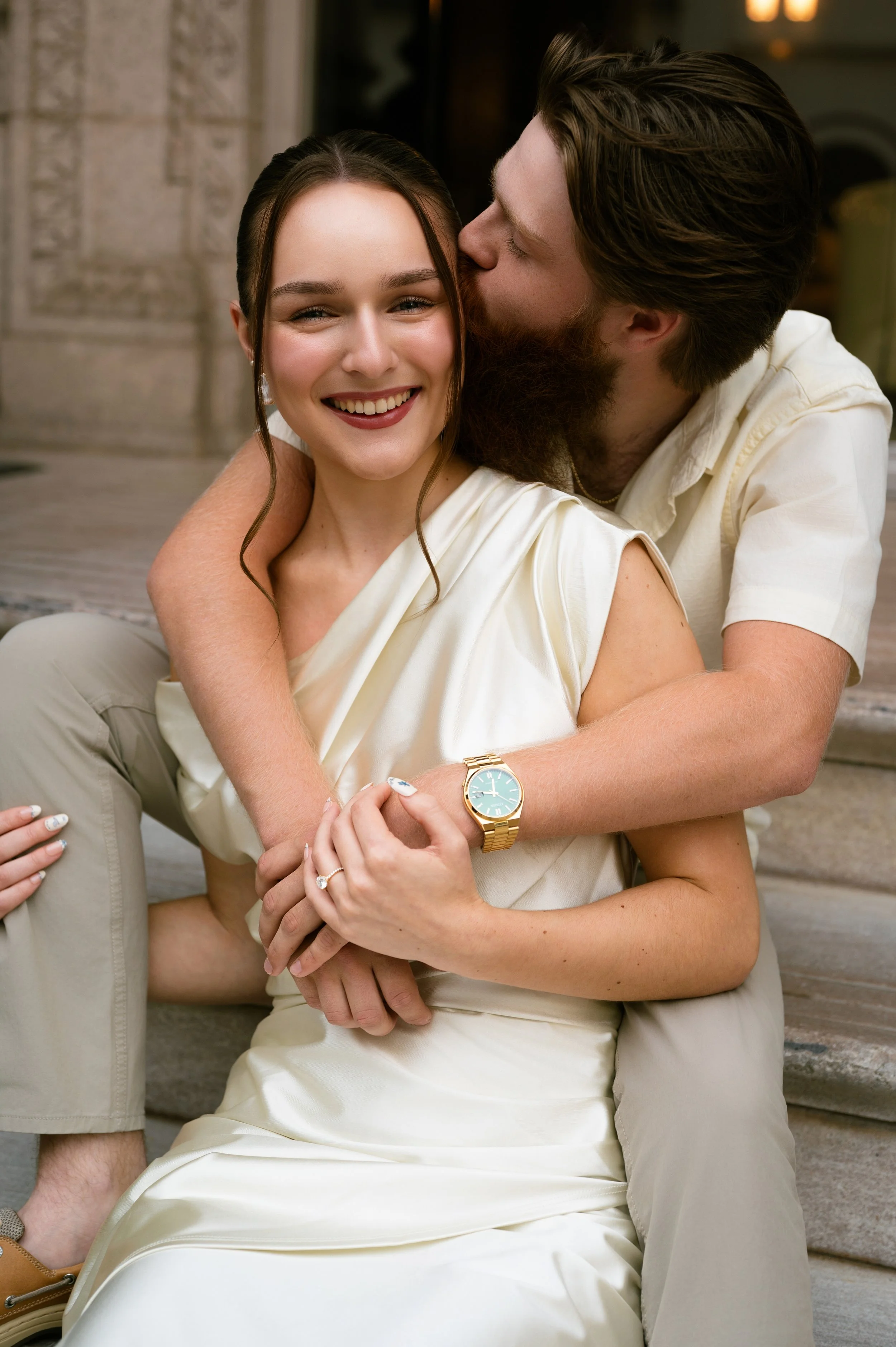 A couple sitting on stairs, with the man kissing the woman's cheek. The woman is smiling, and the man has his arms around her. The woman is wearing a white dress and a gold watch.
