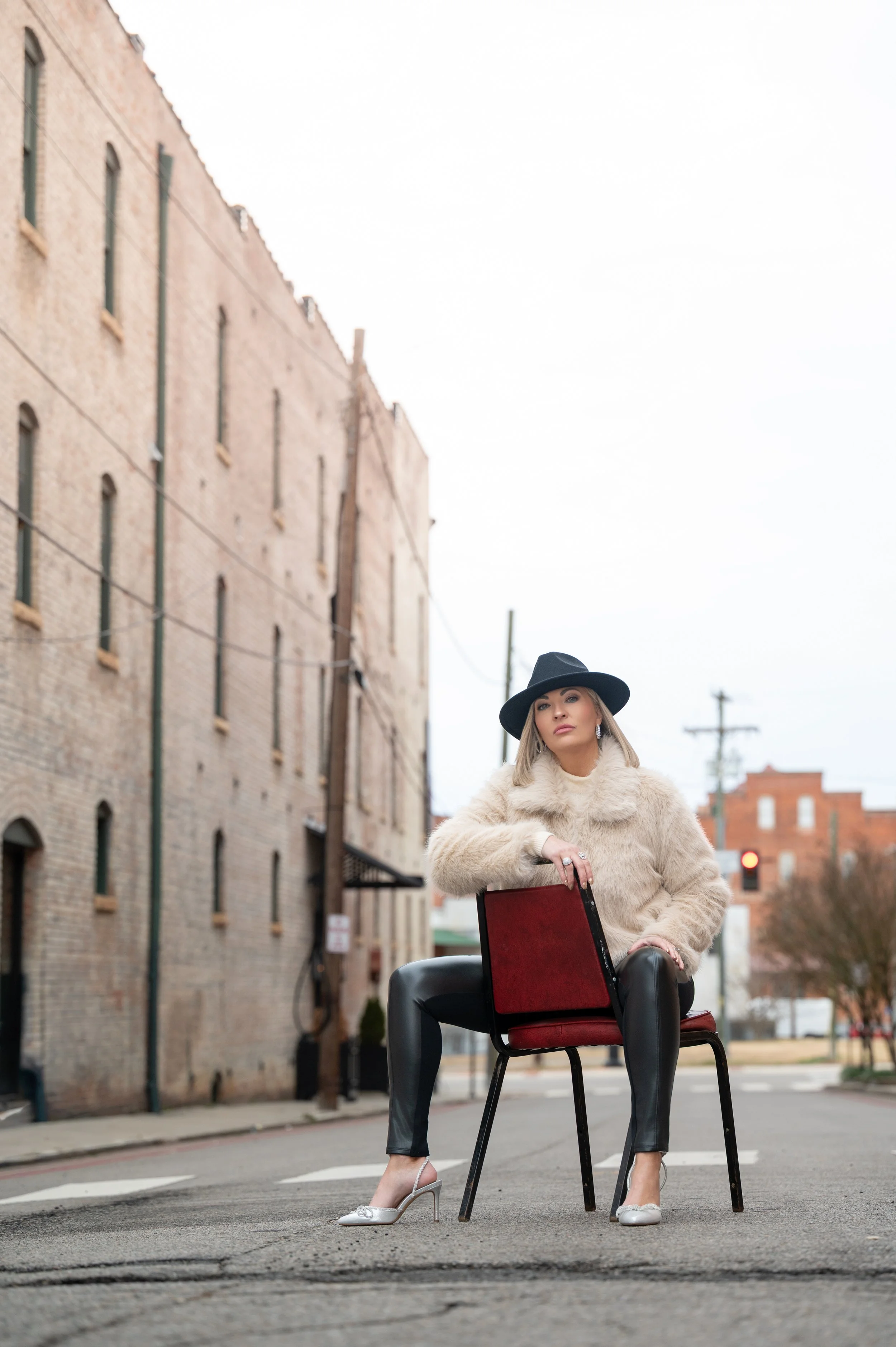 A woman wearing a fur coat, black leather pants, and high heels sitting on a chair in the middle of an empty street with brick buildings in the background.