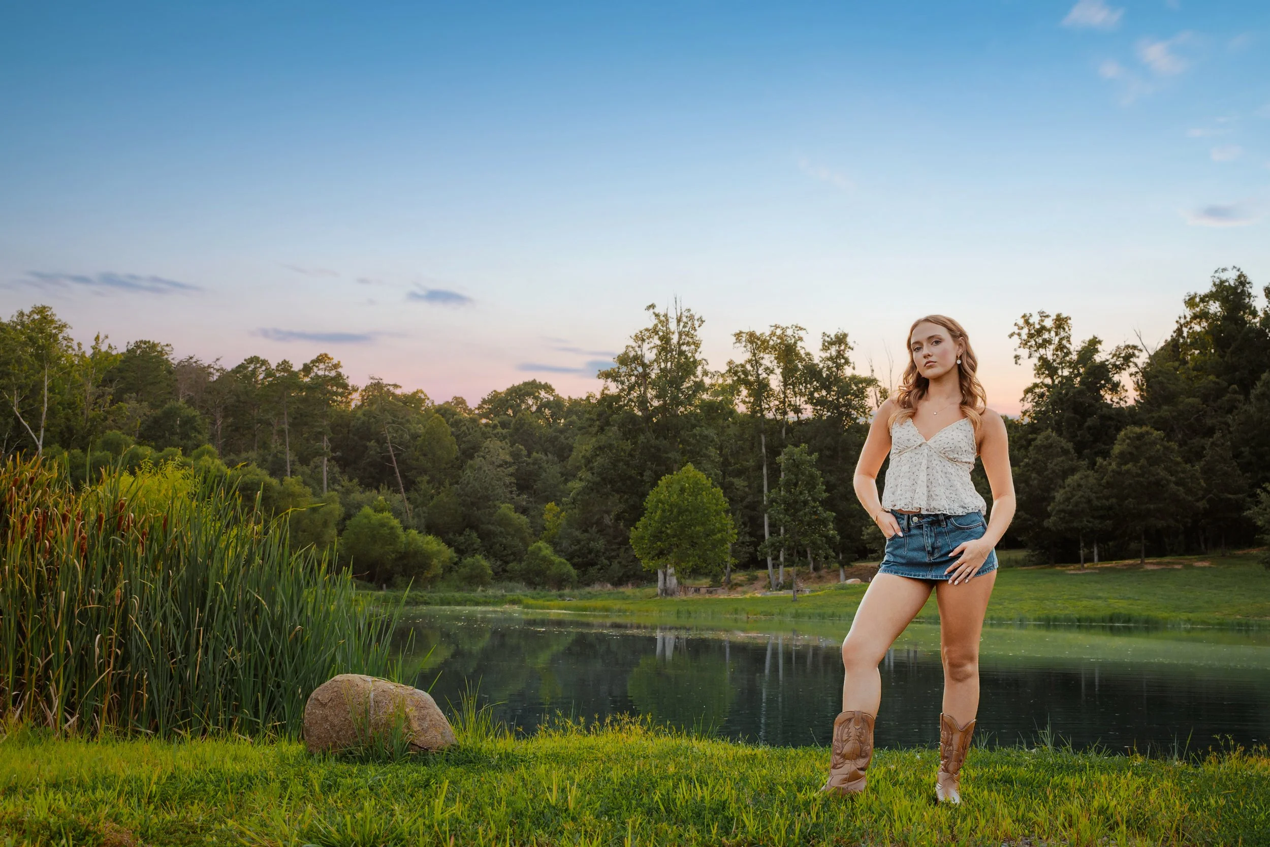 A young woman standing on green grass near a lake with a forest and trees in the background during sunset or sunrise.