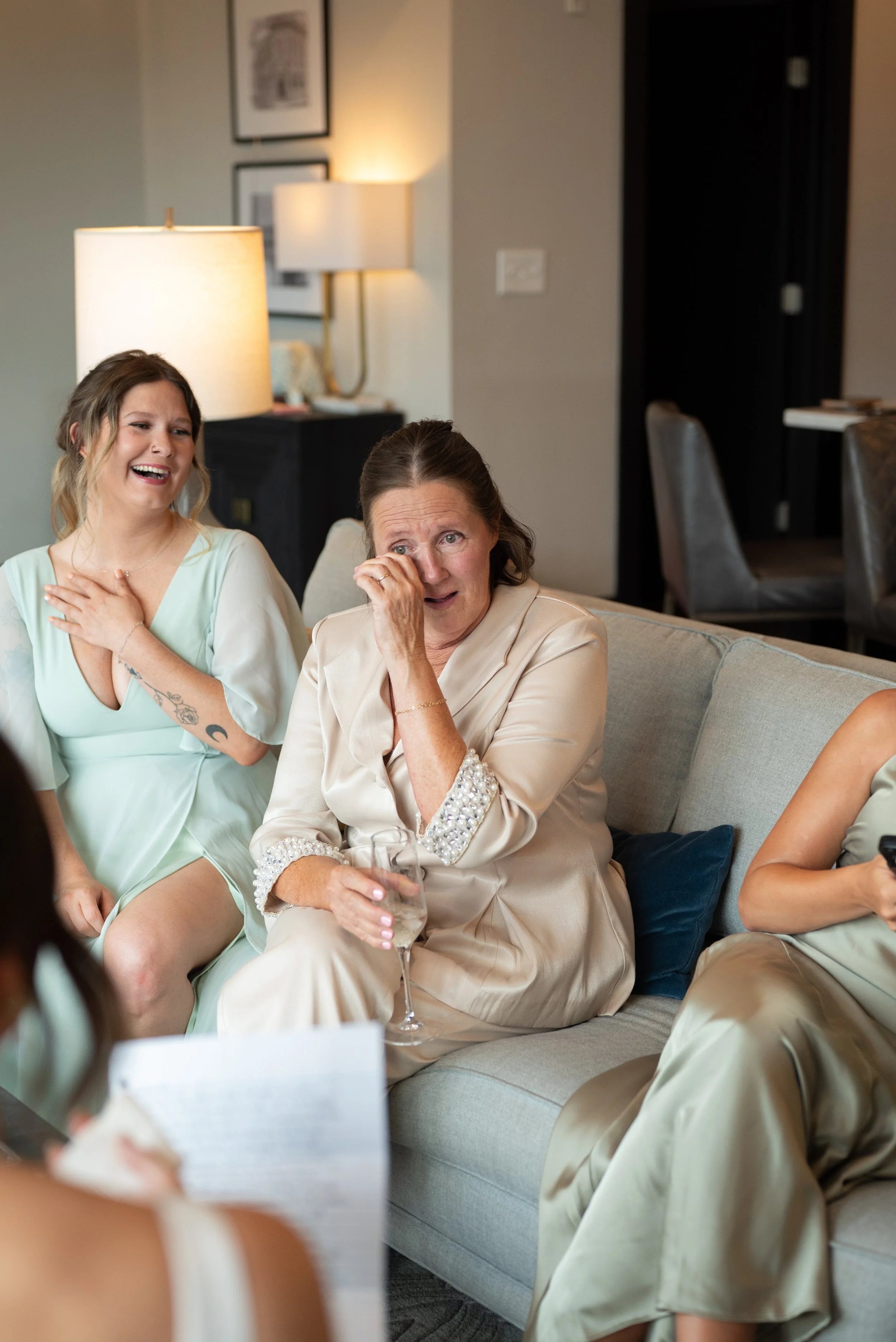 Three women sitting on a sofa, with one woman wiping tears from her eyes and another smiling and laughing beside her, in a warmly lit room.