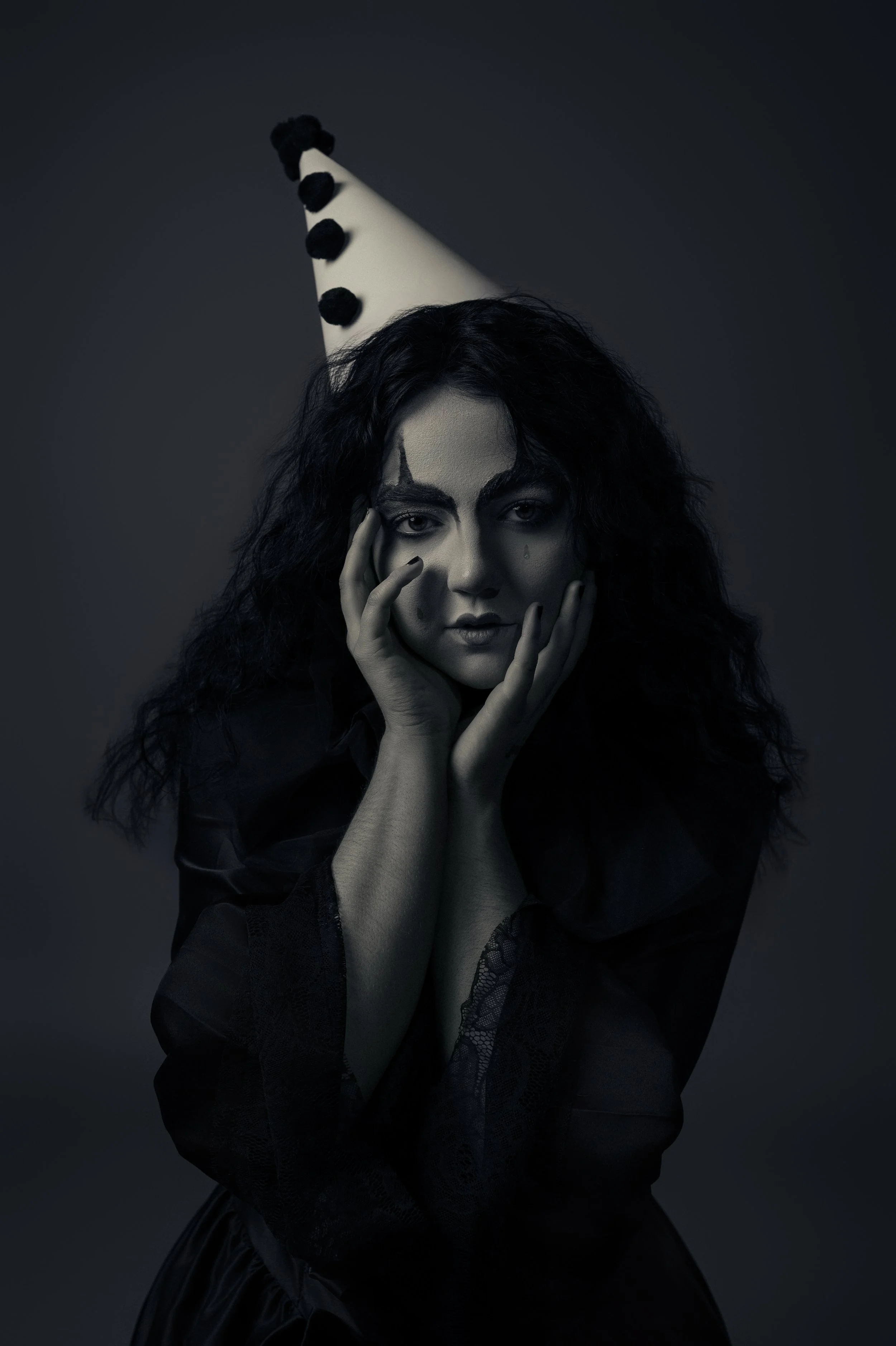 A woman with dark curly hair wearing a black outfit, with her hands on her face, standing against a dark background. She has face makeup resembling a clown, with black lines around her eyes and mouth. She is also wearing a tall, pointed party hat wit