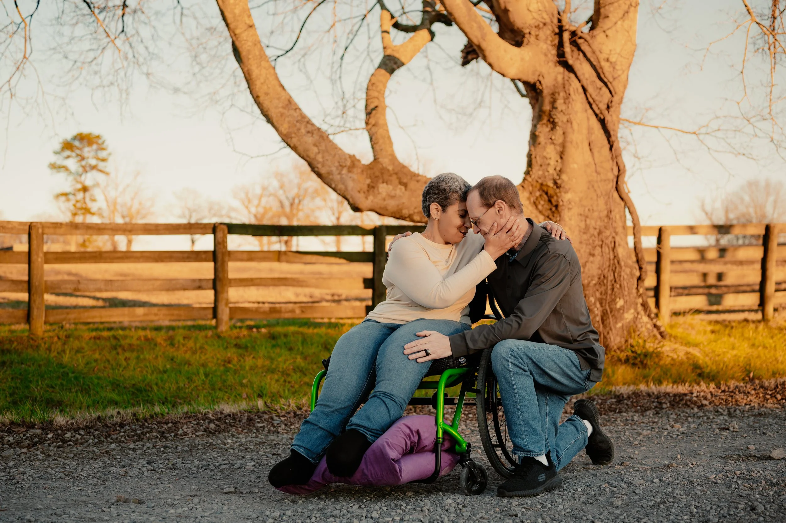 A man and woman sharing a tender moment outdoors beside a large tree, with the woman sitting in a wheelchair and both touching foreheads with eyes closed.