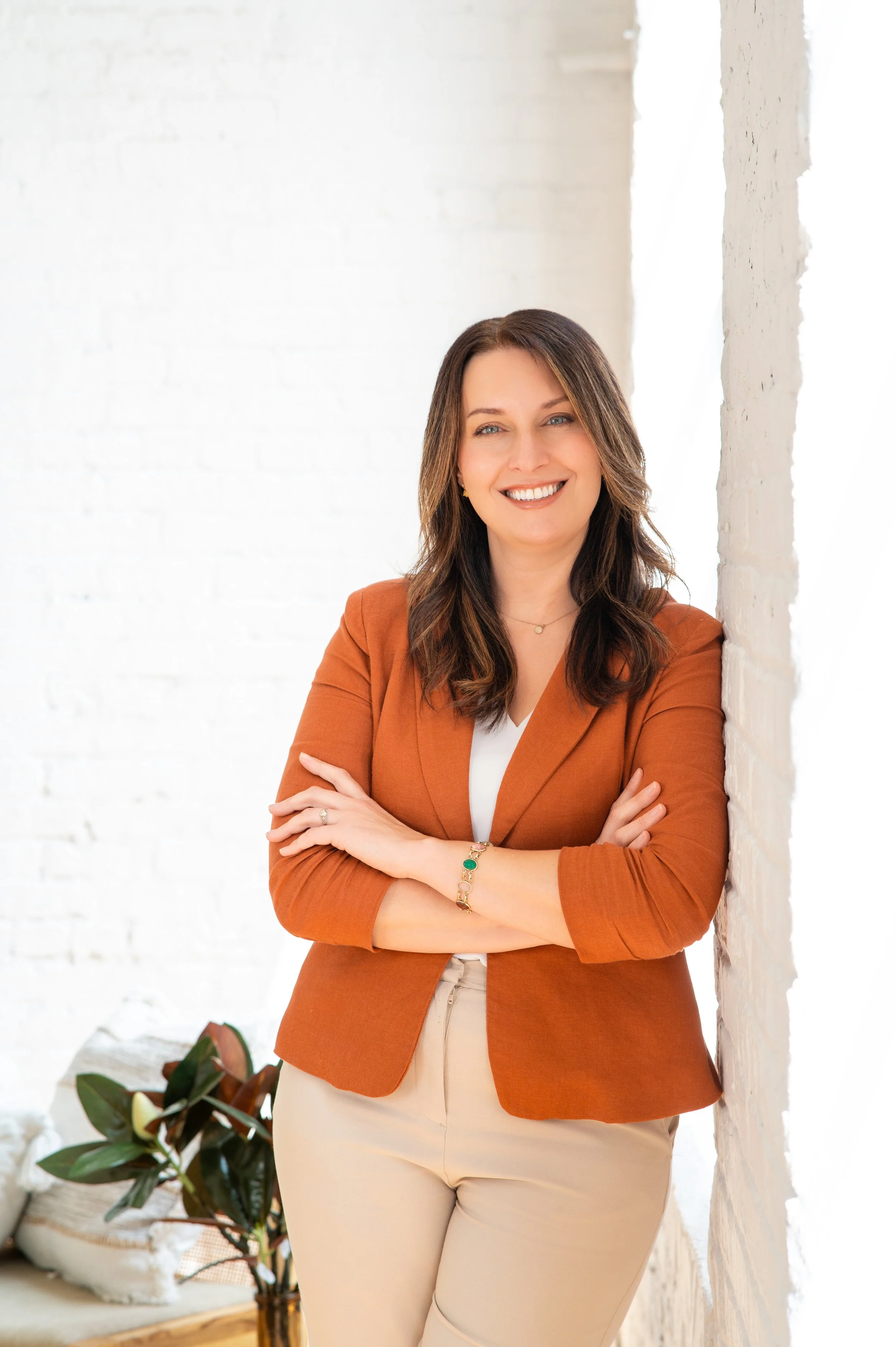 A woman smiling with arms crossed, standing beside a white brick wall in a bright room. headshot studio