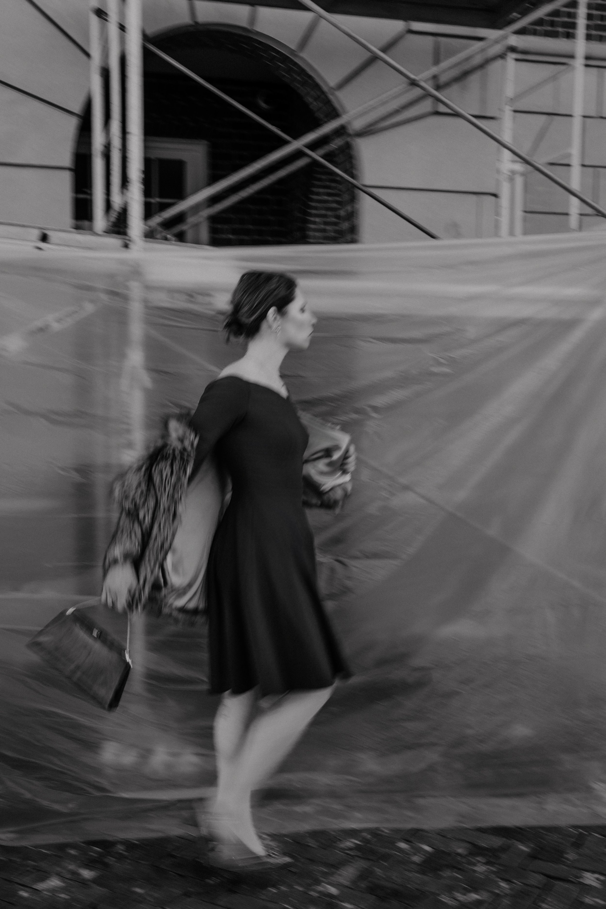 A woman in a black dress walking with a large handbag and coat, outside near a building with scaffolding, in a black-and-white photo.