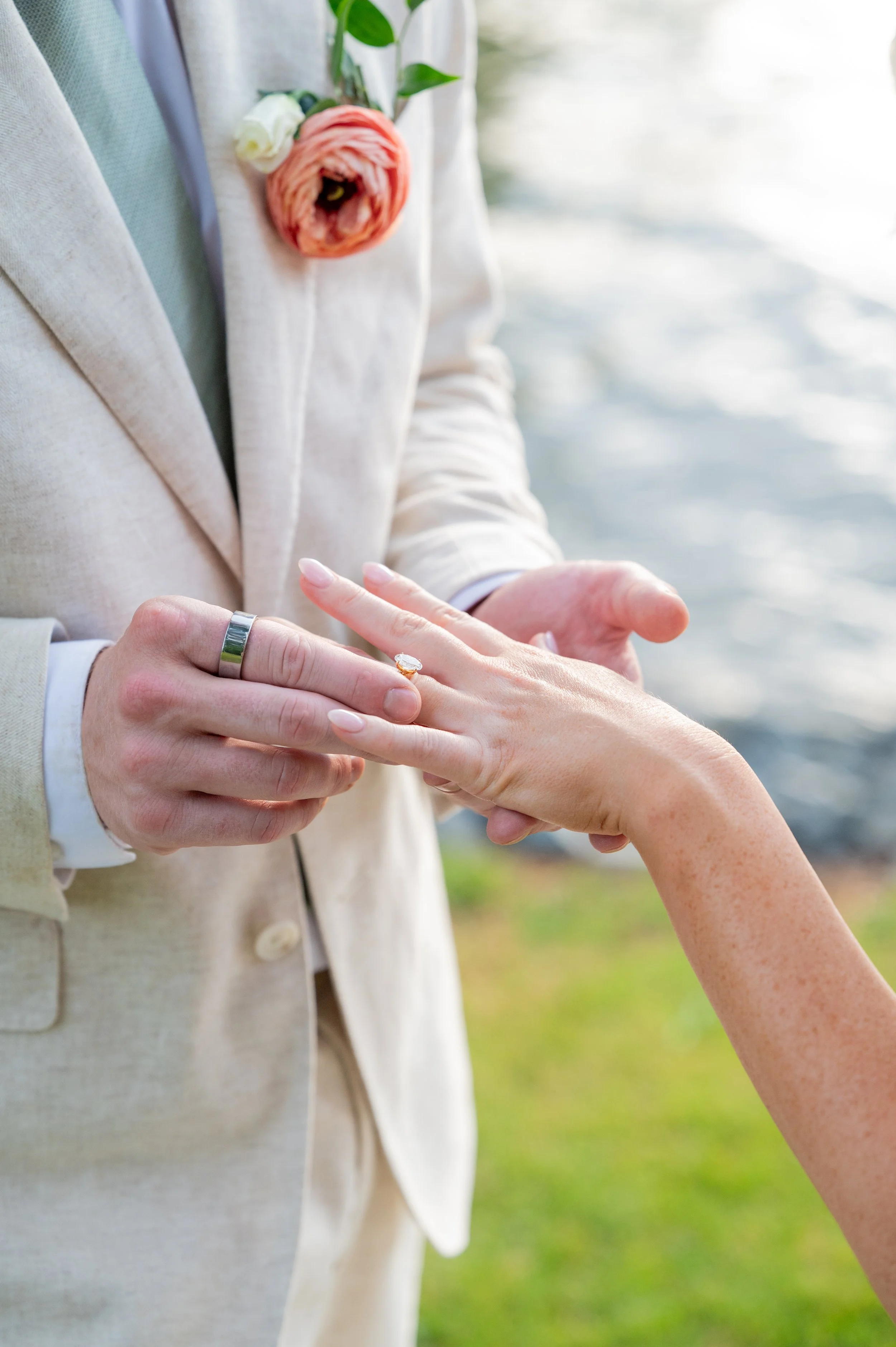 A person in a beige suit is placing a ring on the ring finger of another person's hand, outside near a body of water.