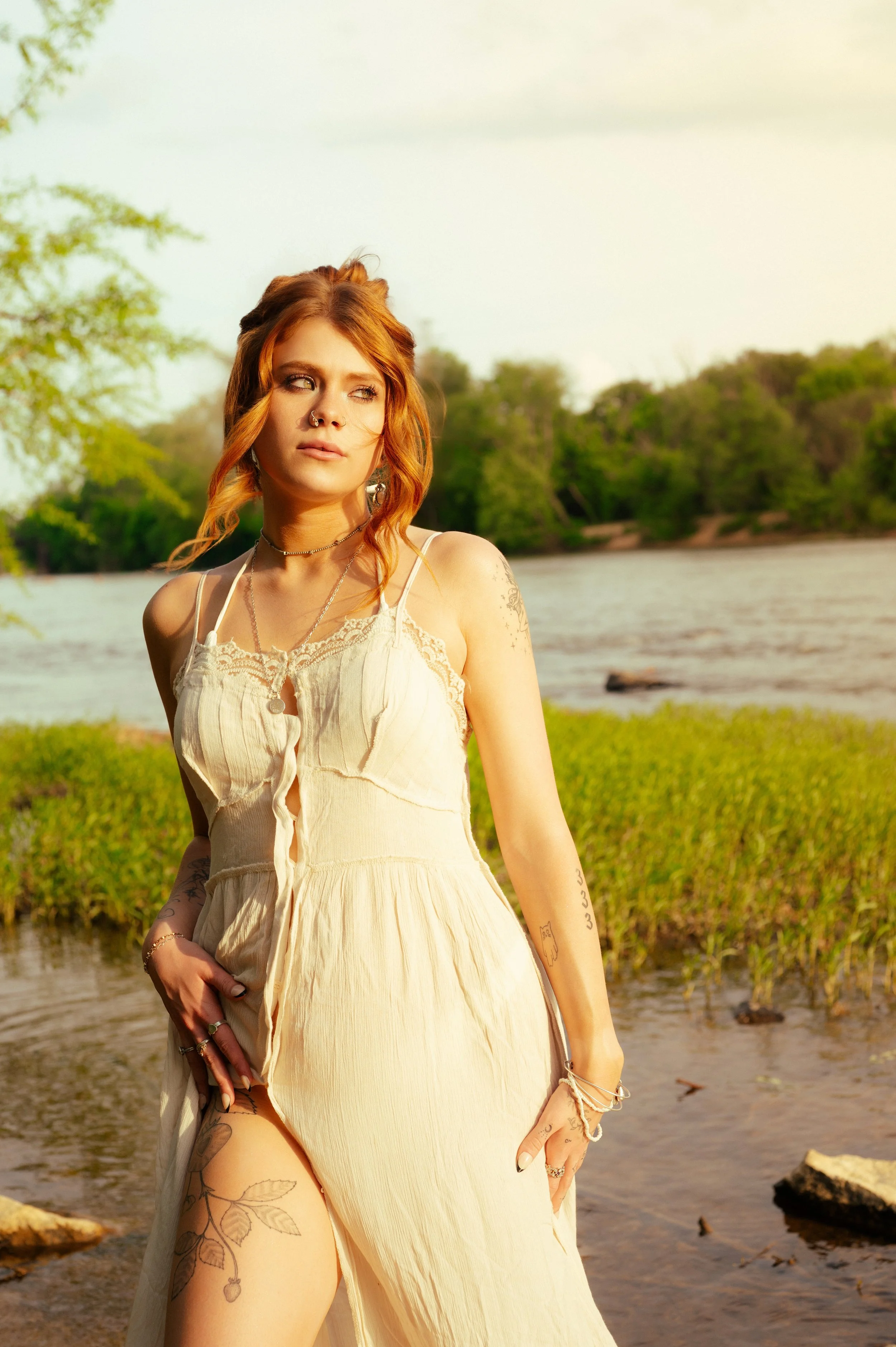 A woman with red hair and tattoos stands near a riverbank, wearing a white dress with lace details, looking to her right with a contemplative expression, during daytime.