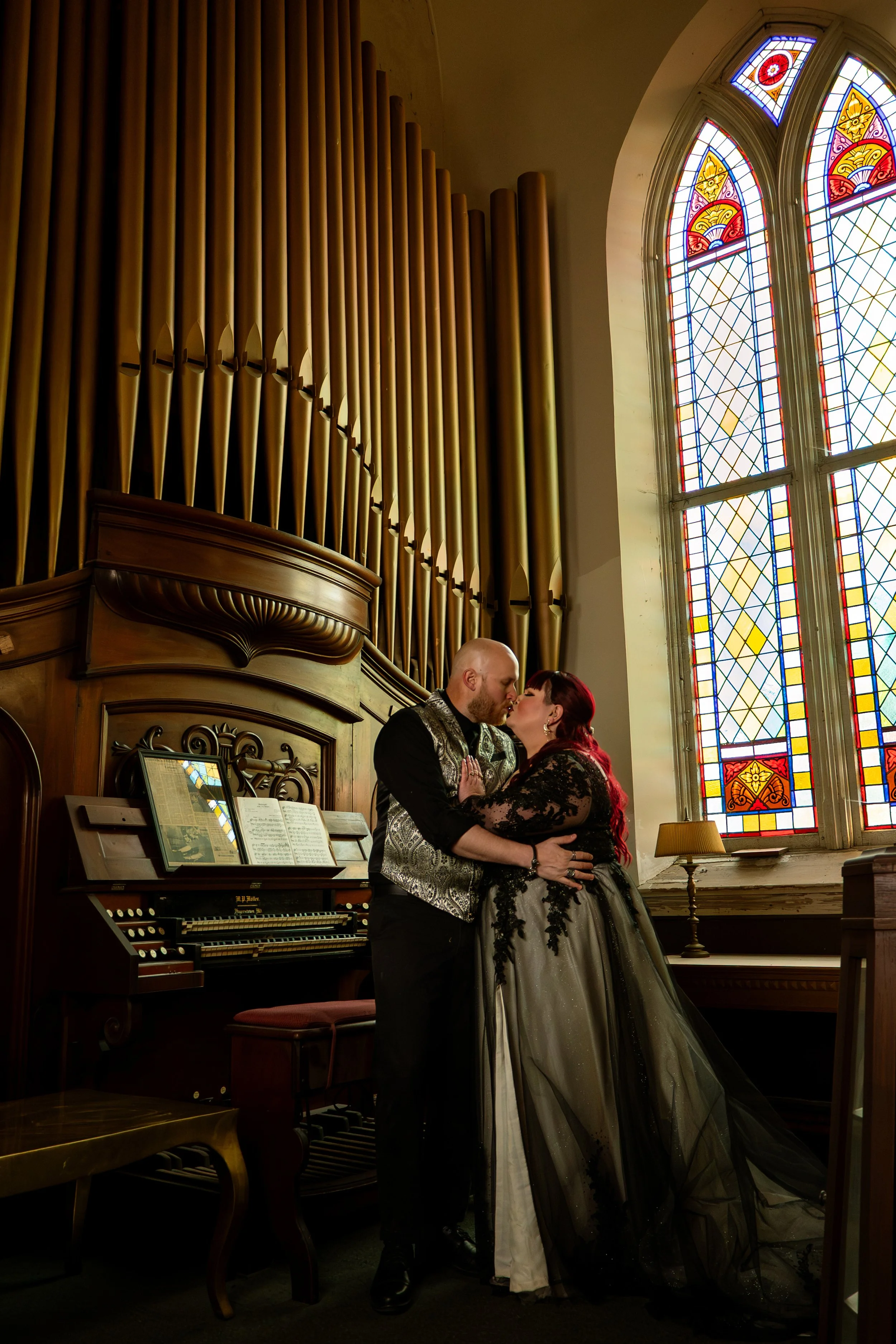 A couple sharing a kiss inside a church, near a stained glass window with organ pipes in the background.