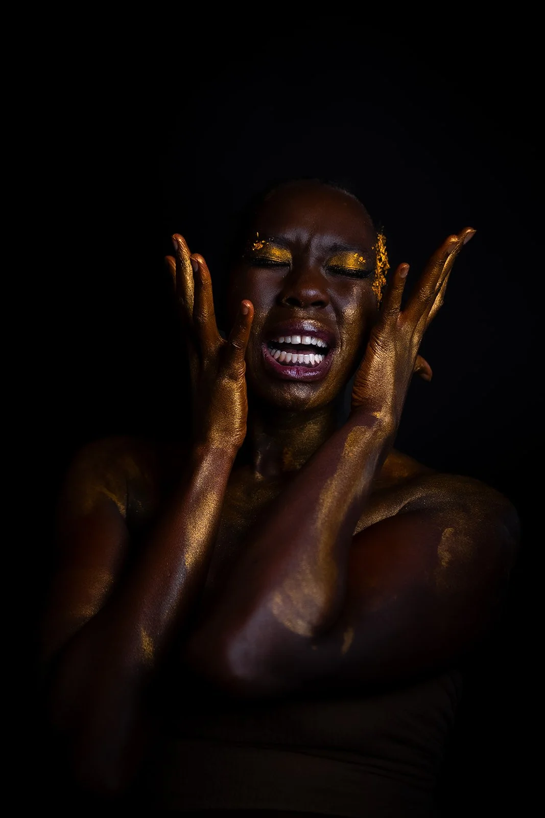 A woman with gold paint on her face and body, with her eyes closed and mouth open, touching her face with both hands against a dark background.