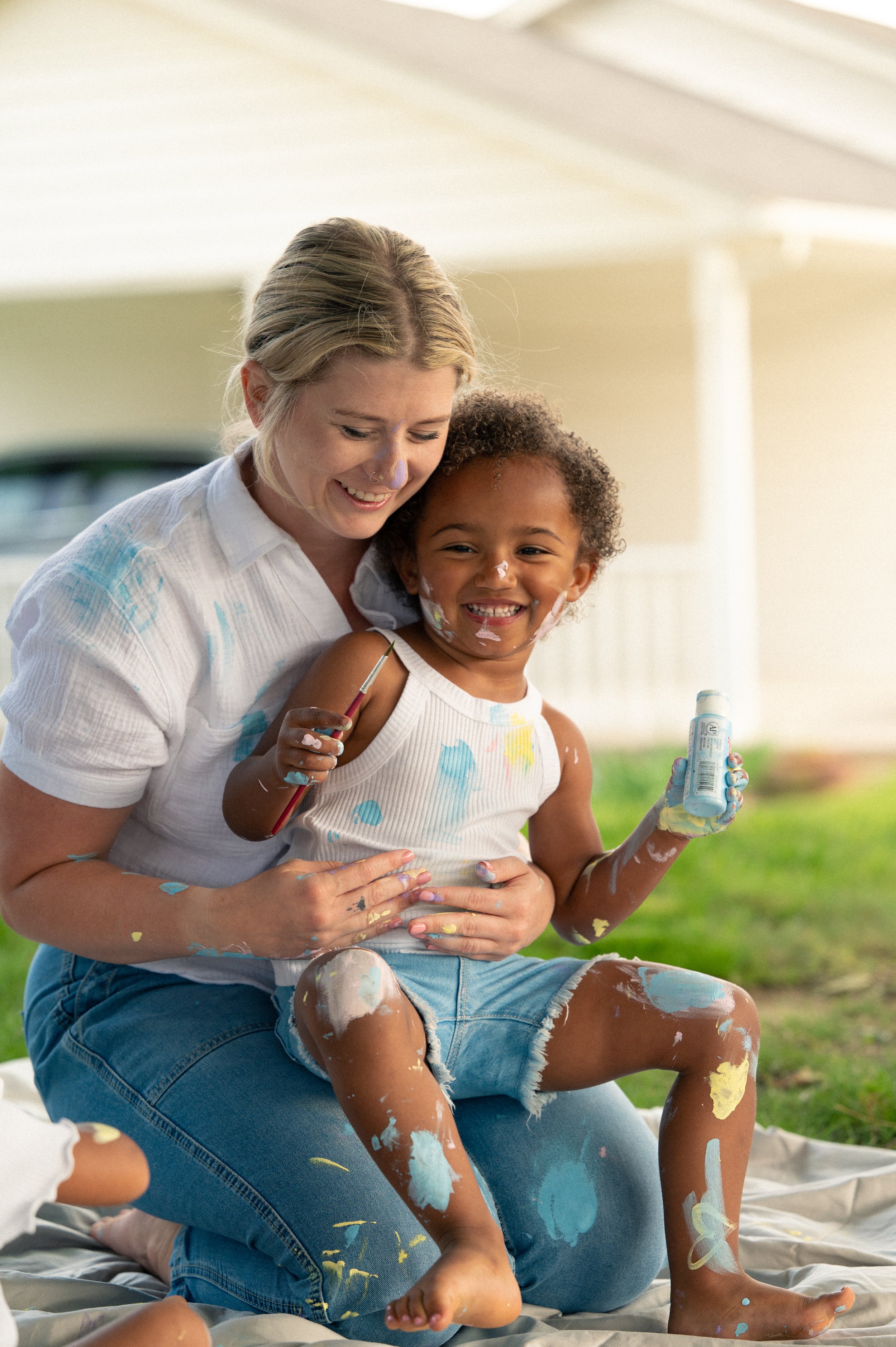 A woman and young girl playing with paint outside, covered in multicolored paint spots, sitting on a blanket and smiling.