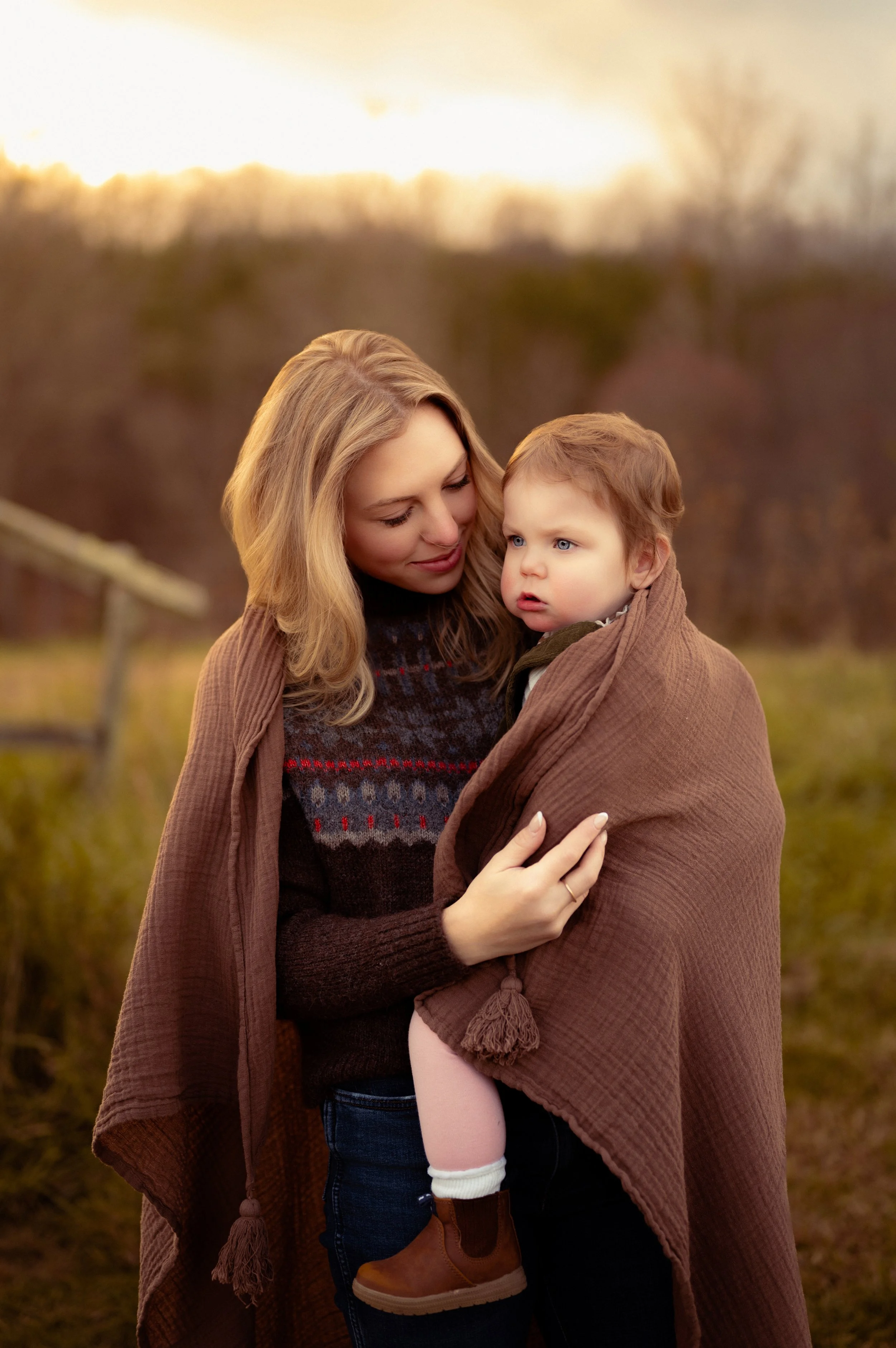 A woman holding a young child wrapped in a brown blanket outdoors during sunset.