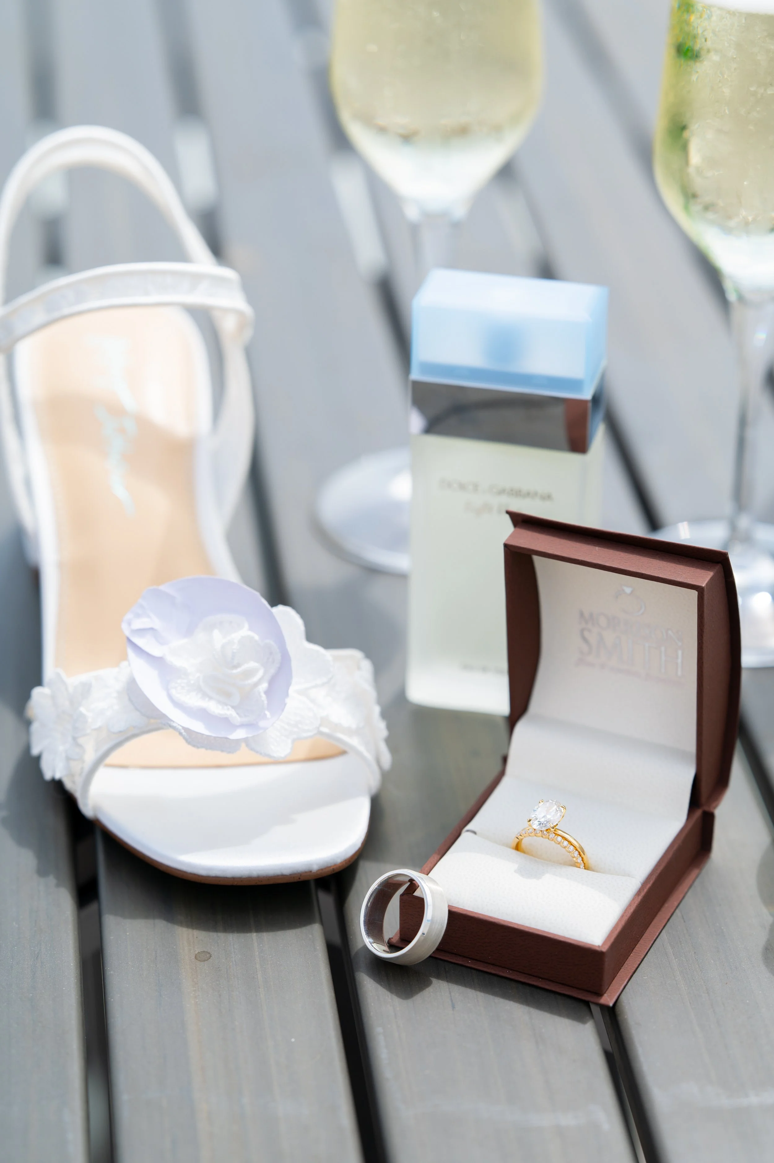 Wedding details with bride’s shoe, rings, and perfume on a table in natural light.
