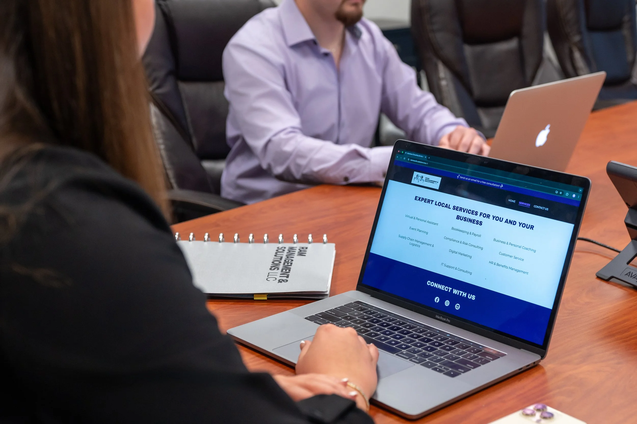 Person working on a laptop at a business meeting, with another person in the background also working on a laptop.
