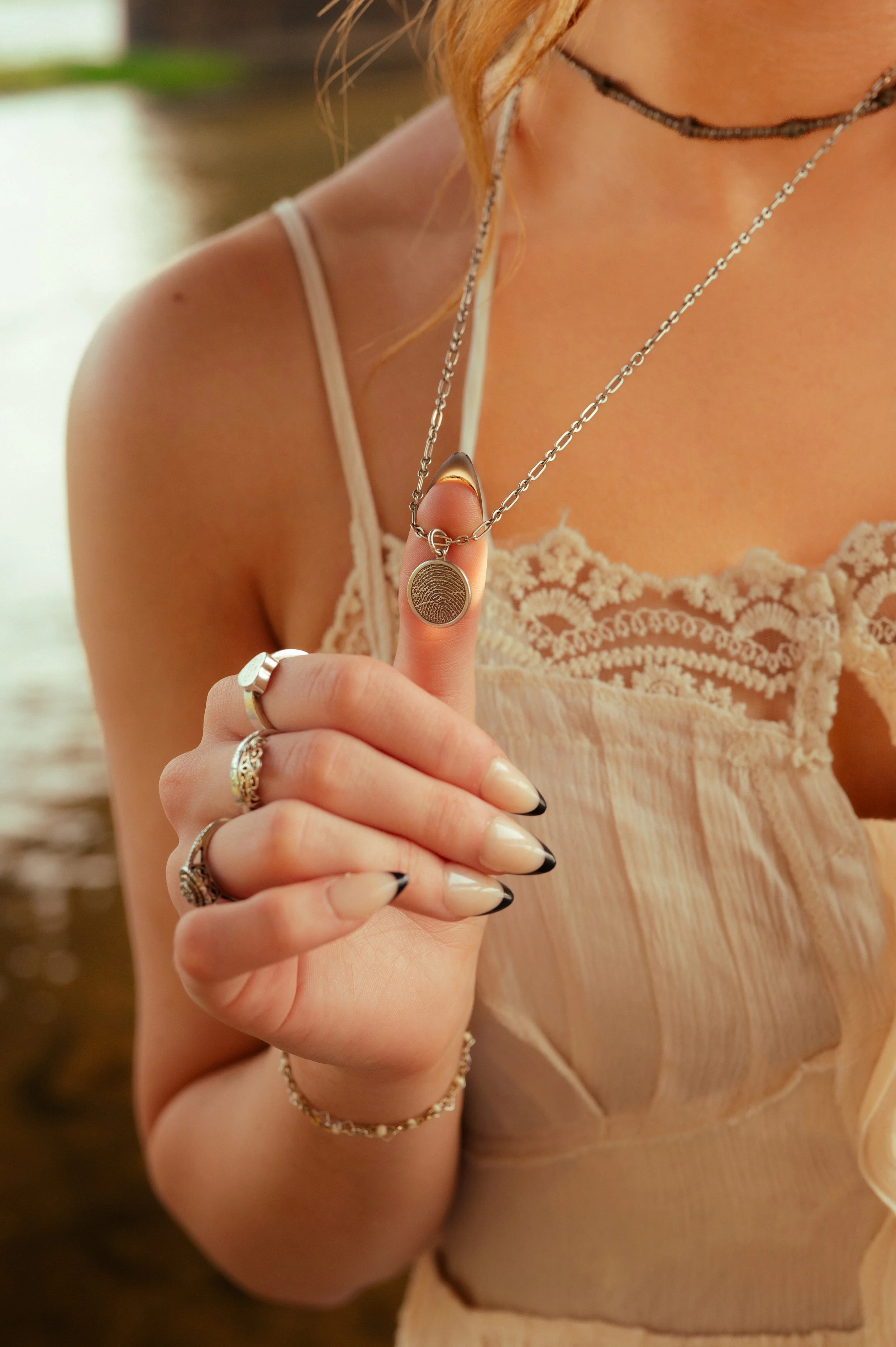 Close-up of a woman holding a fingerprint-themed pendant necklace, with her fingernails painted black with natural tips, wearing multiple rings and a lace-trimmed beige dress.