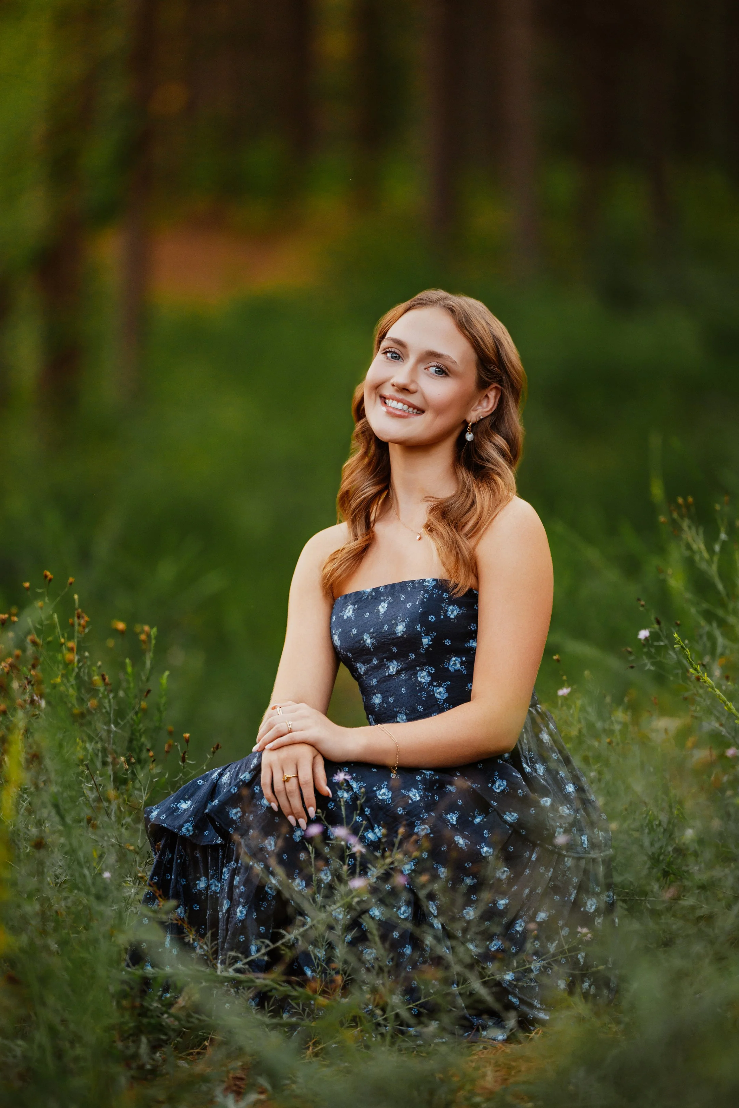 A young woman with long, wavy, red hair and blue eyes sitting on grass in a field of wildflowers, smiling at the camera, wearing a dark blue strapless dress with a floral pattern.