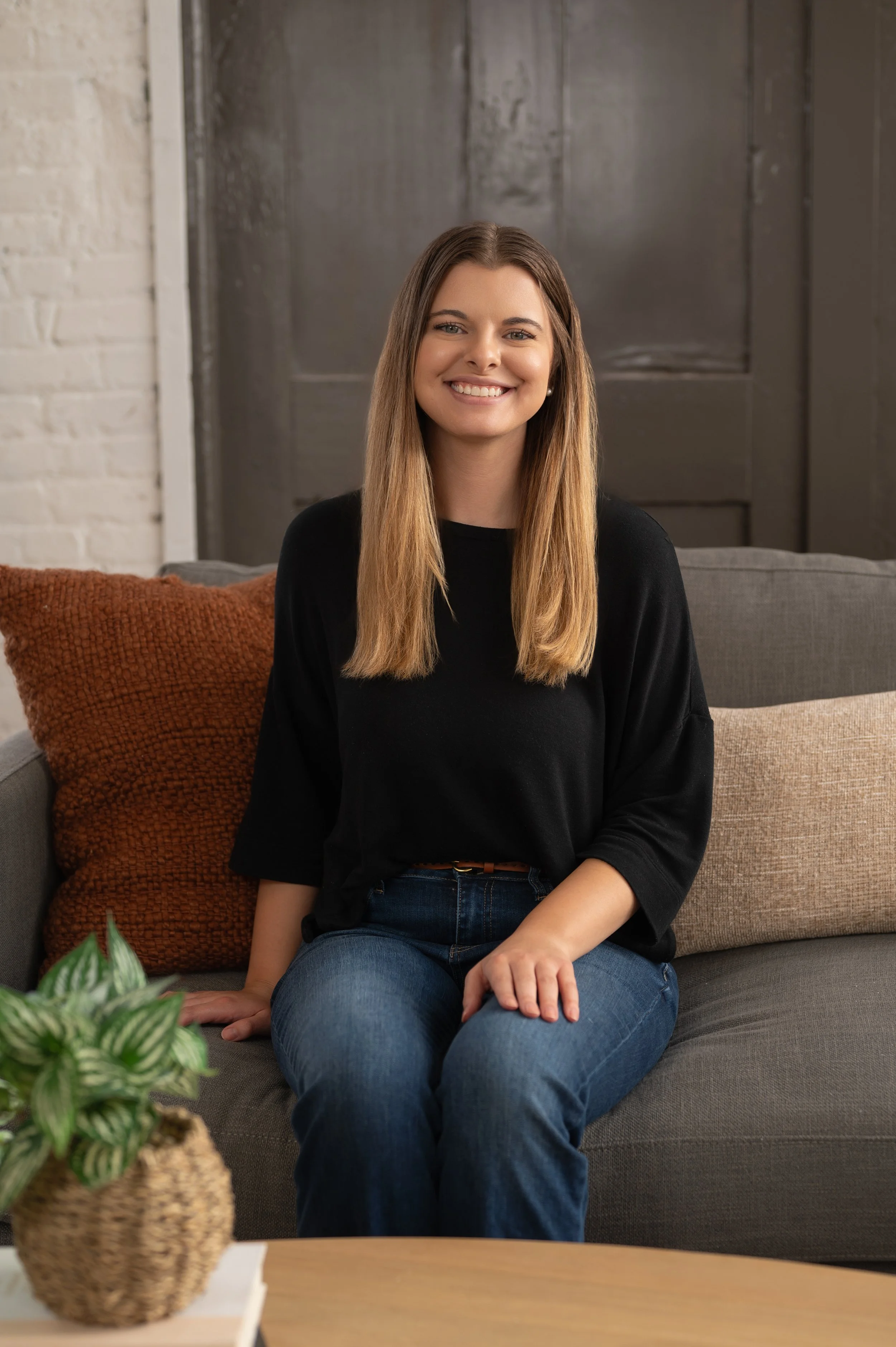 A young woman with long, straight light brown hair, smiling while sitting on a gray sofa. She is wearing a black top and blue jeans. There are two pillows behind her, one orange and one beige, and a potted plant on a table in front of her.