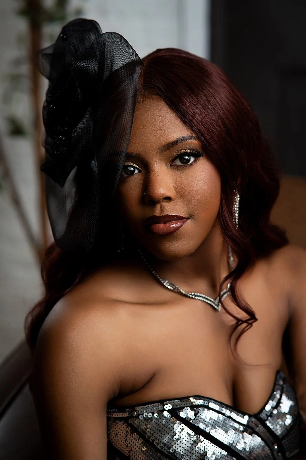 A woman with dark skin and wavy auburn hair wearing a strapless sequined dress, a black fascinator with netting, earrings, and a necklace, looking directly at the camera.