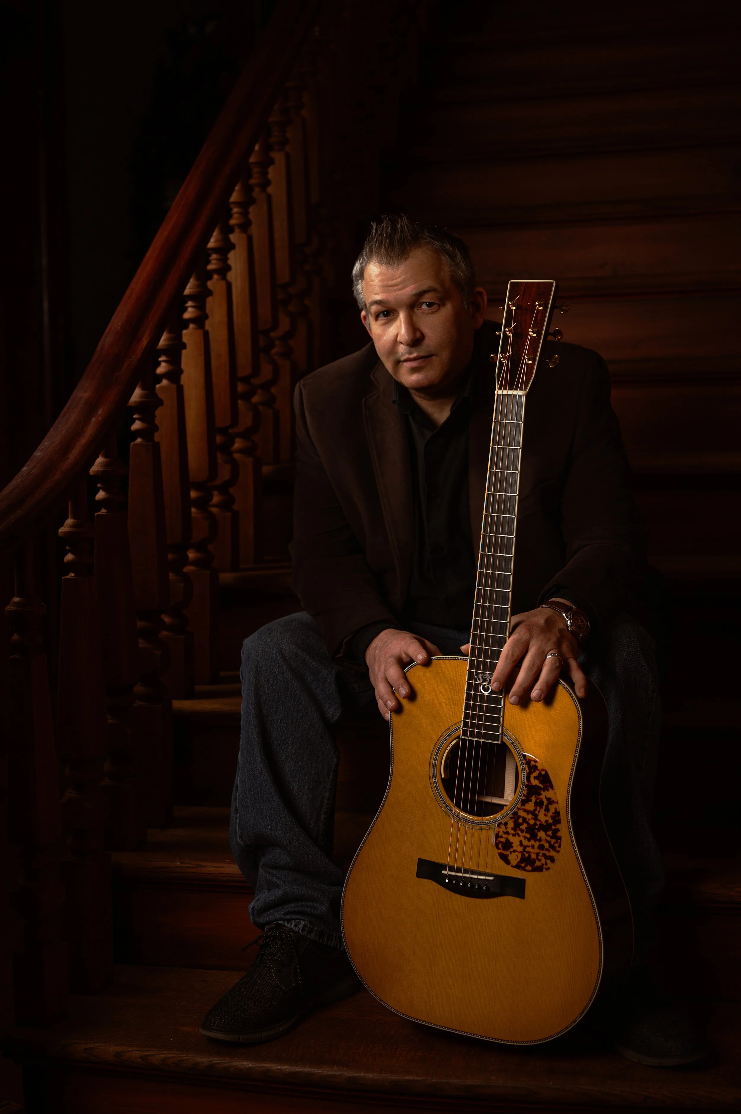 A man sitting on wooden stairs holding an acoustic guitar, with a dark wooden staircase in the background.