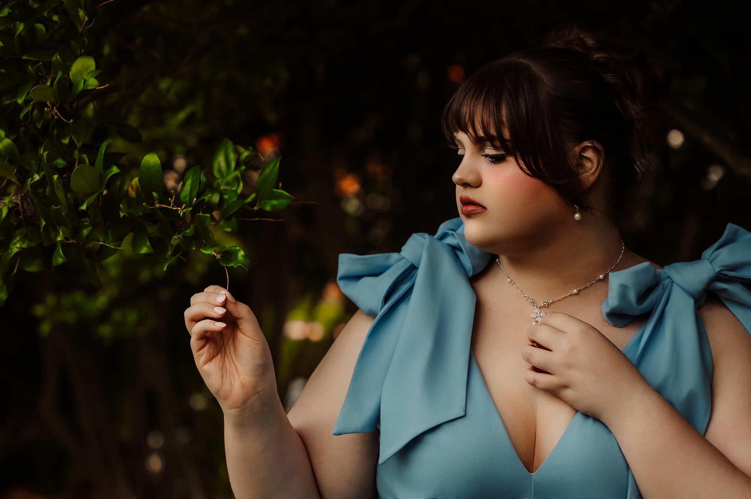 A woman in a light blue dress with shoulder bows looking at a branch with green leaves in a dark outdoor setting, wearing jewelry including earrings, a necklace, and a ring.