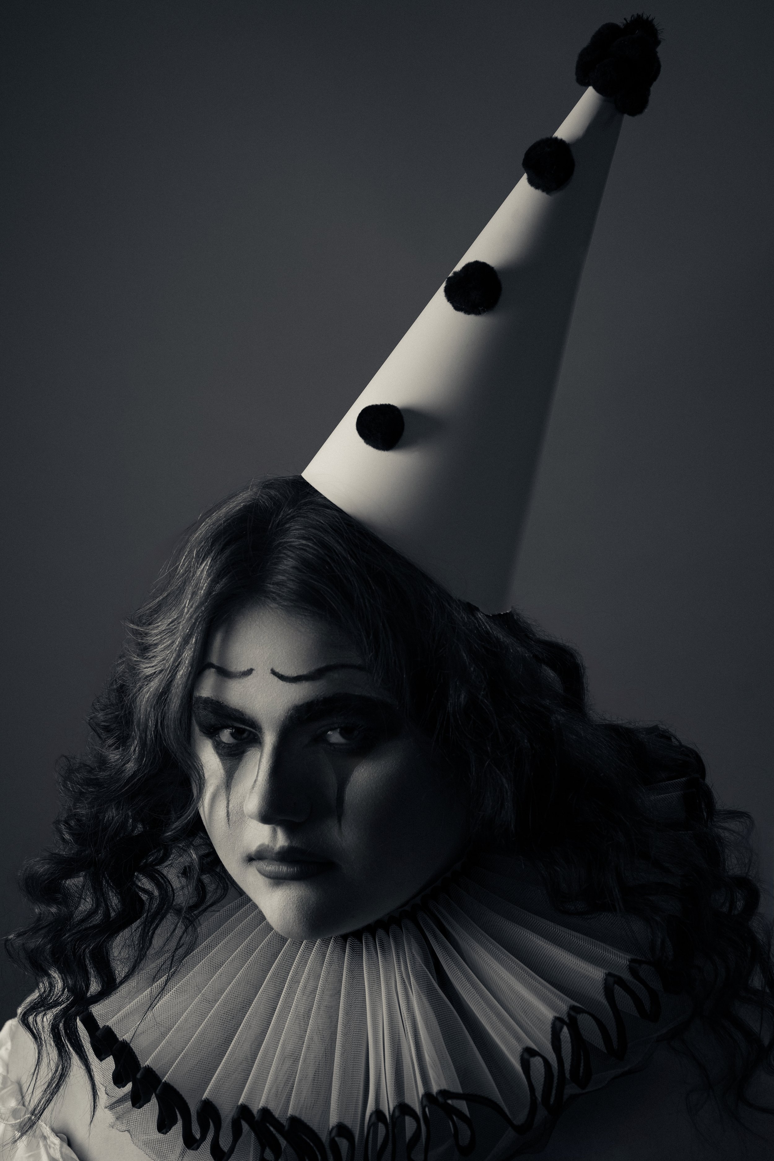 A woman with clown makeup and curly hair wearing a traditional clown ruffle collar and a tall birthday hat with pom-poms, in a black and white photo.