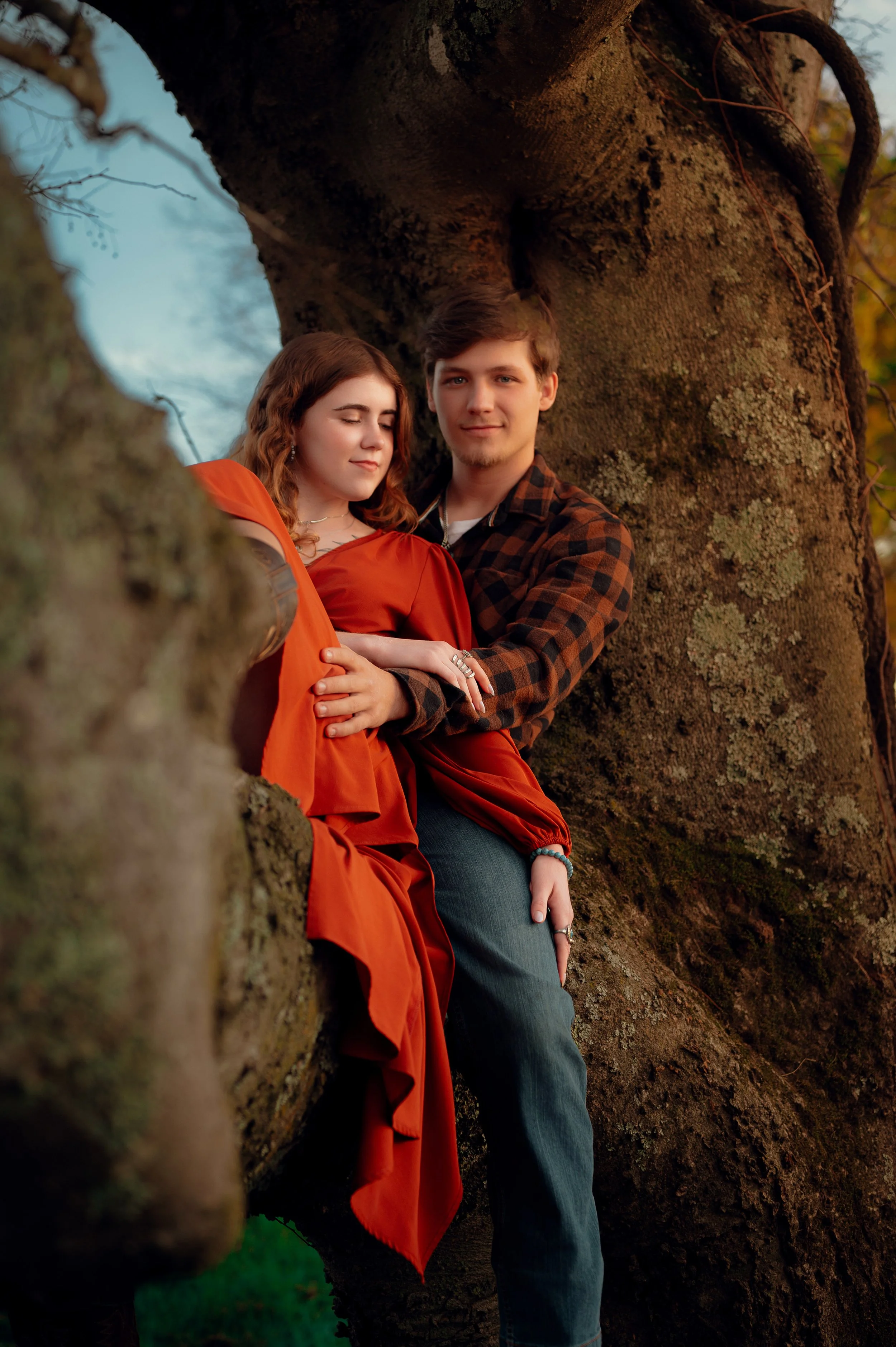 A couple sitting on a tree branch during sunset, with the woman wearing an orange dress and the man wearing a checkered shirt, embracing against a large tree trunk.