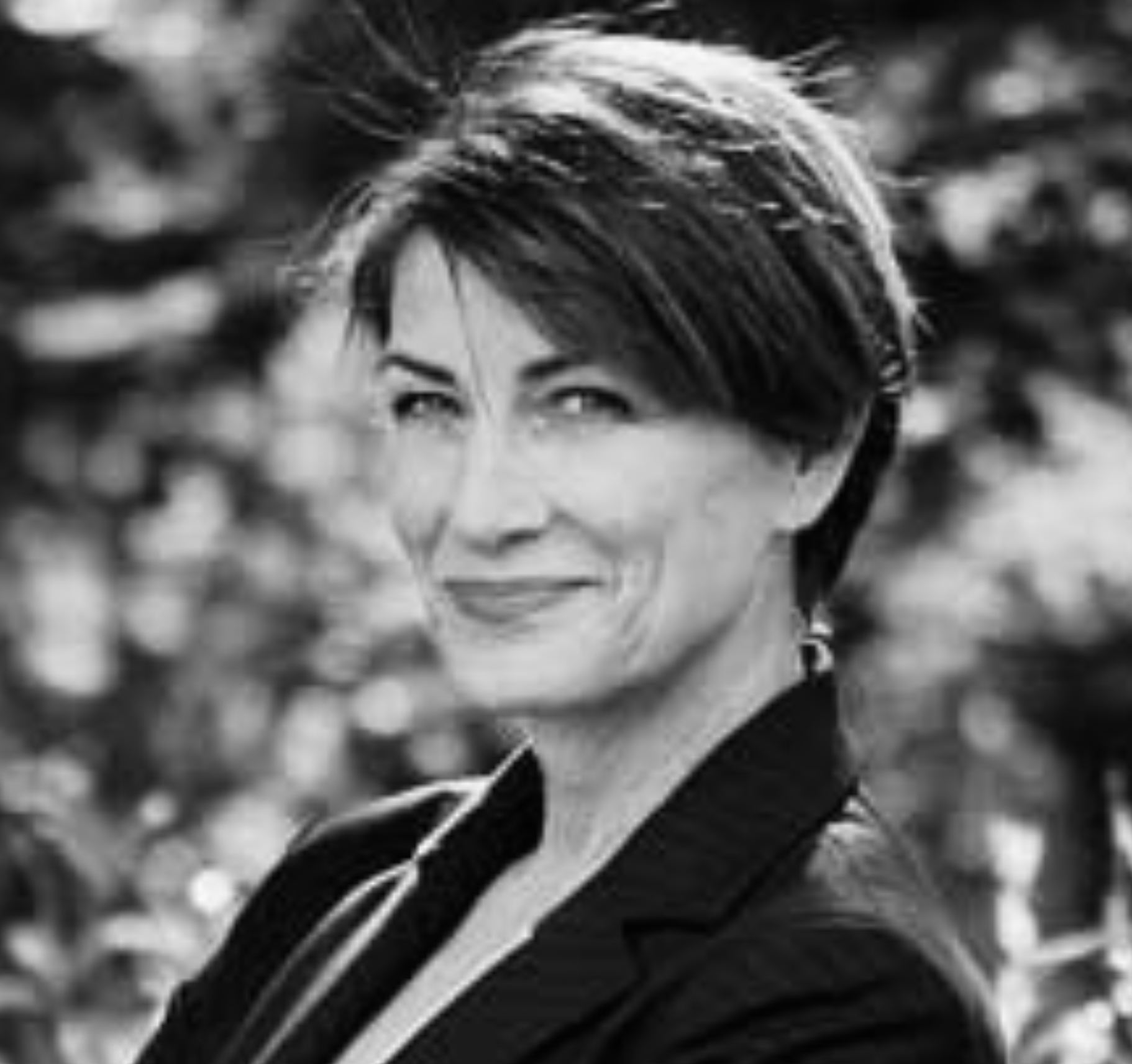A woman with short dark hair and earrings standing amidst blurred foliage or trees.