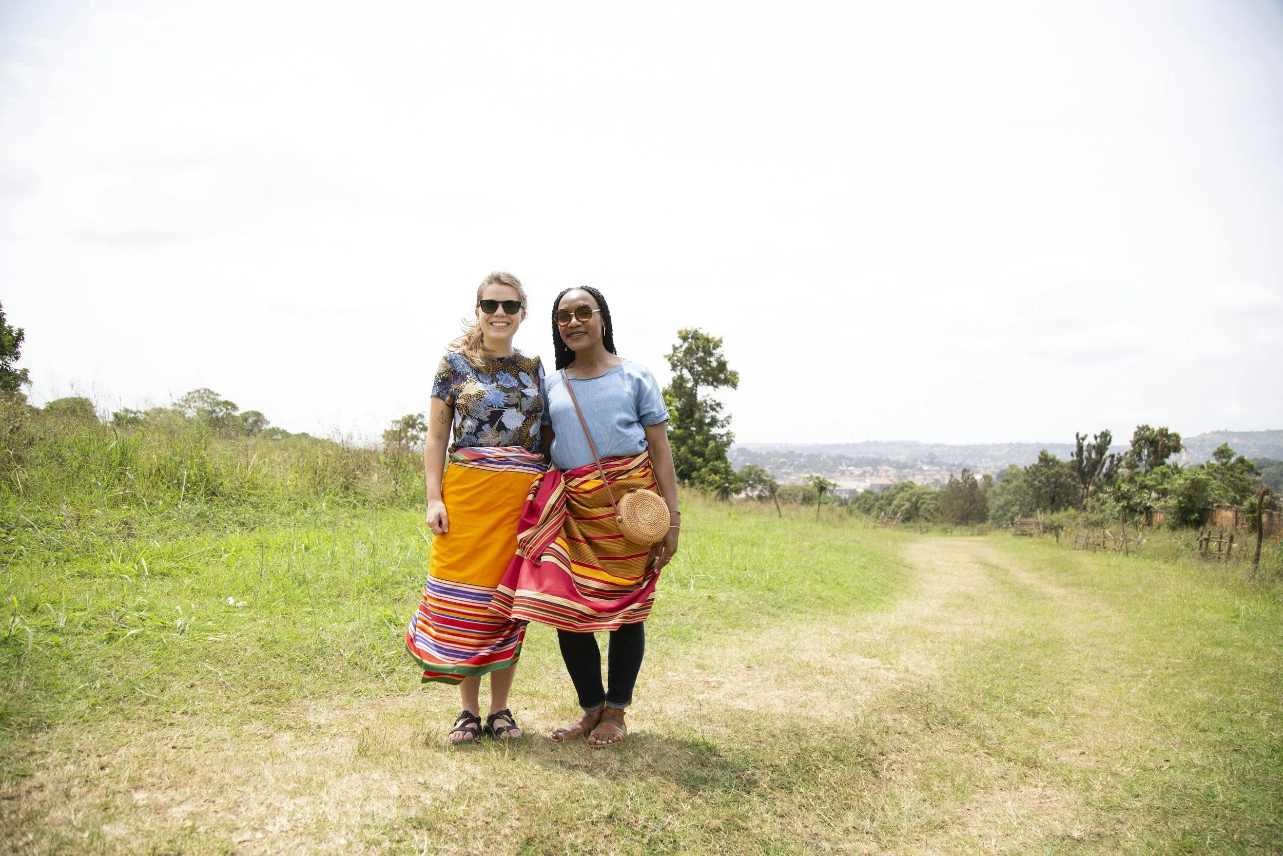 Two women standing on a grassy field, wearing colorful traditional skirts, sunglasses, and smiling.