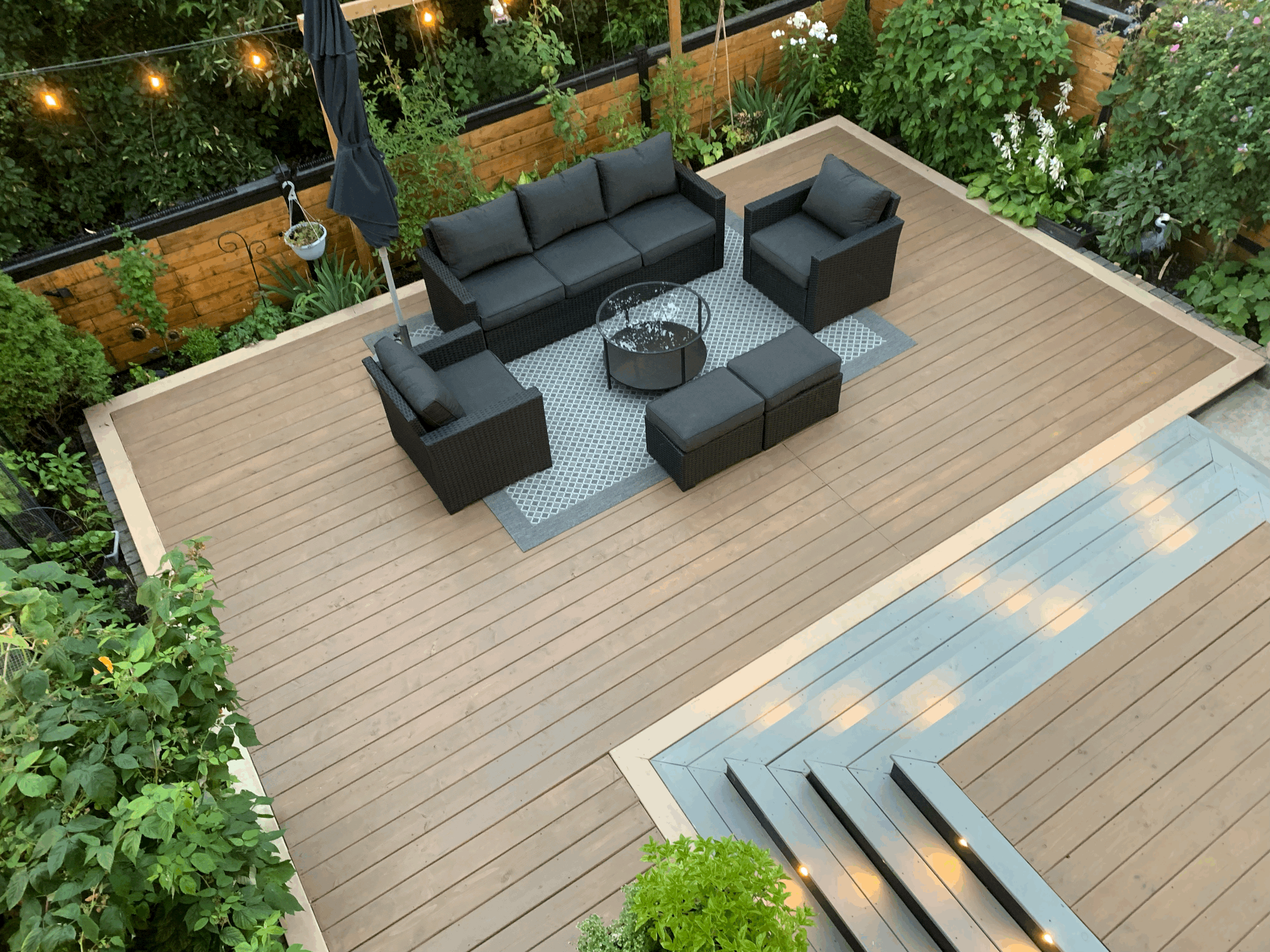 Outdoor wooden deck area with a black outdoor sofa set, a patterned outdoor rug, and surrounding greenery and string lights.