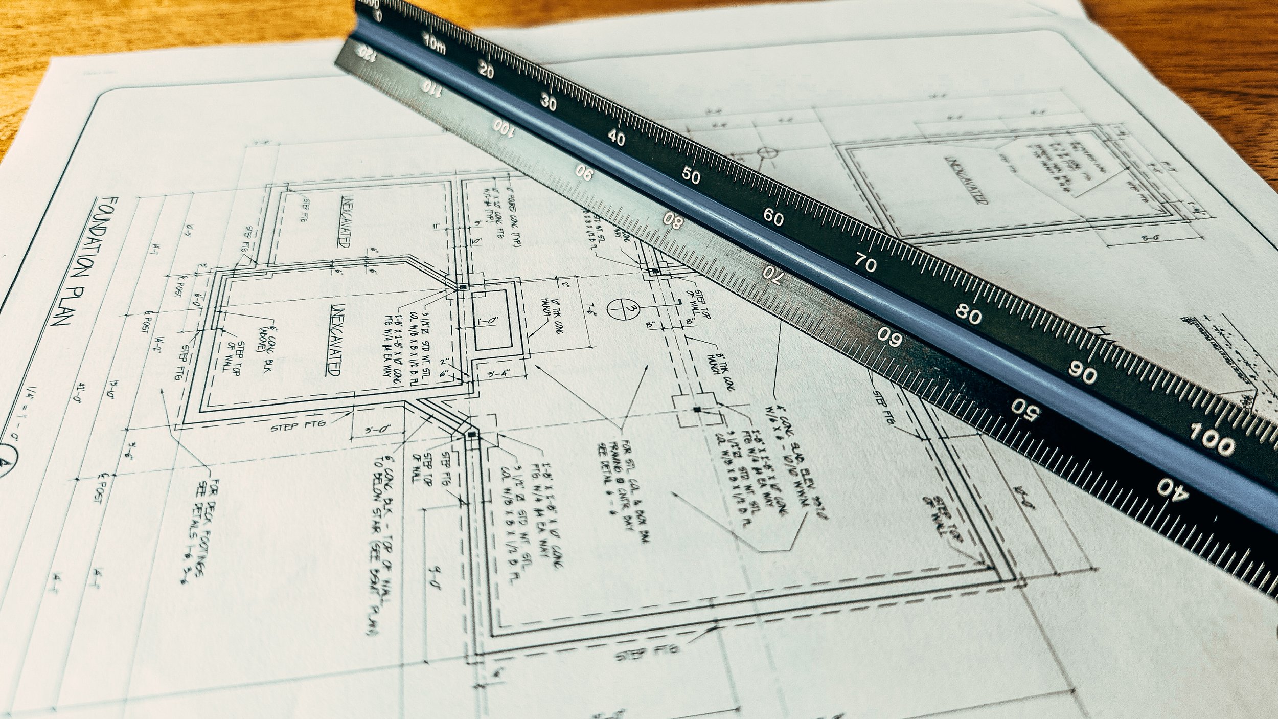 Architectural blueprint with detailed measurements and notes on a foundation plan, overlaid with a metal ruler.