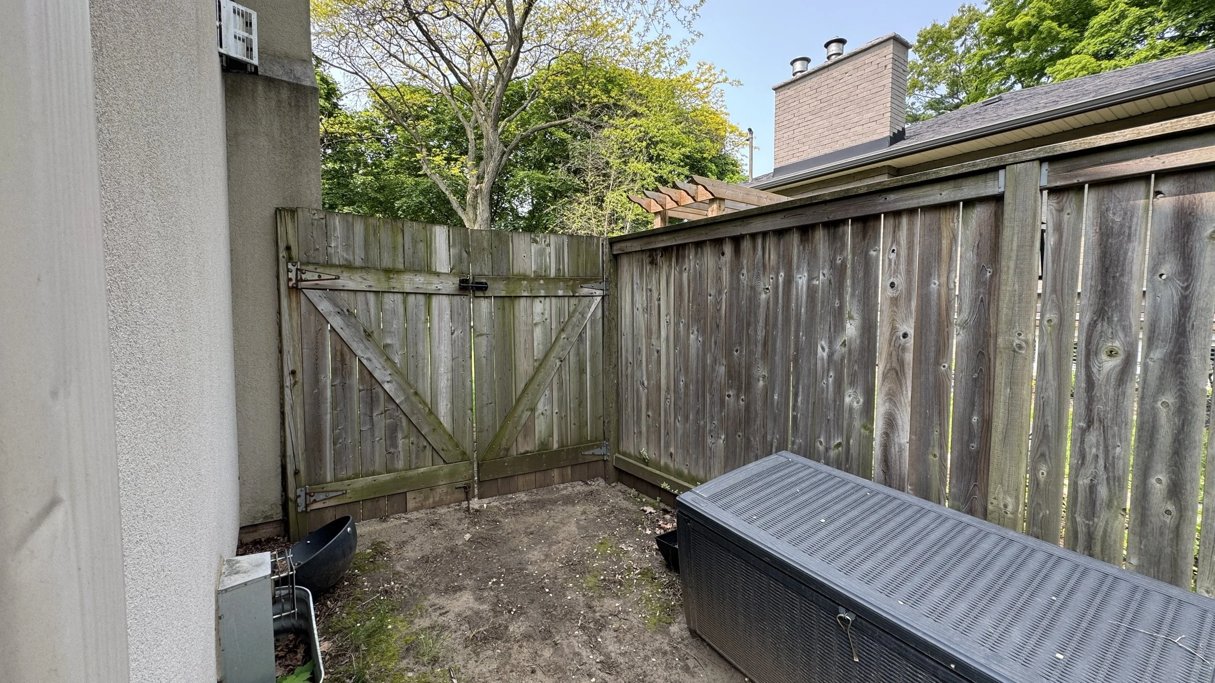 Small backyard with a dirt ground, wooden fence, a wooden gate, and a gray storage chest. There is a black container and some gardening tools on the left side near the house wall. Green trees are visible in the background.