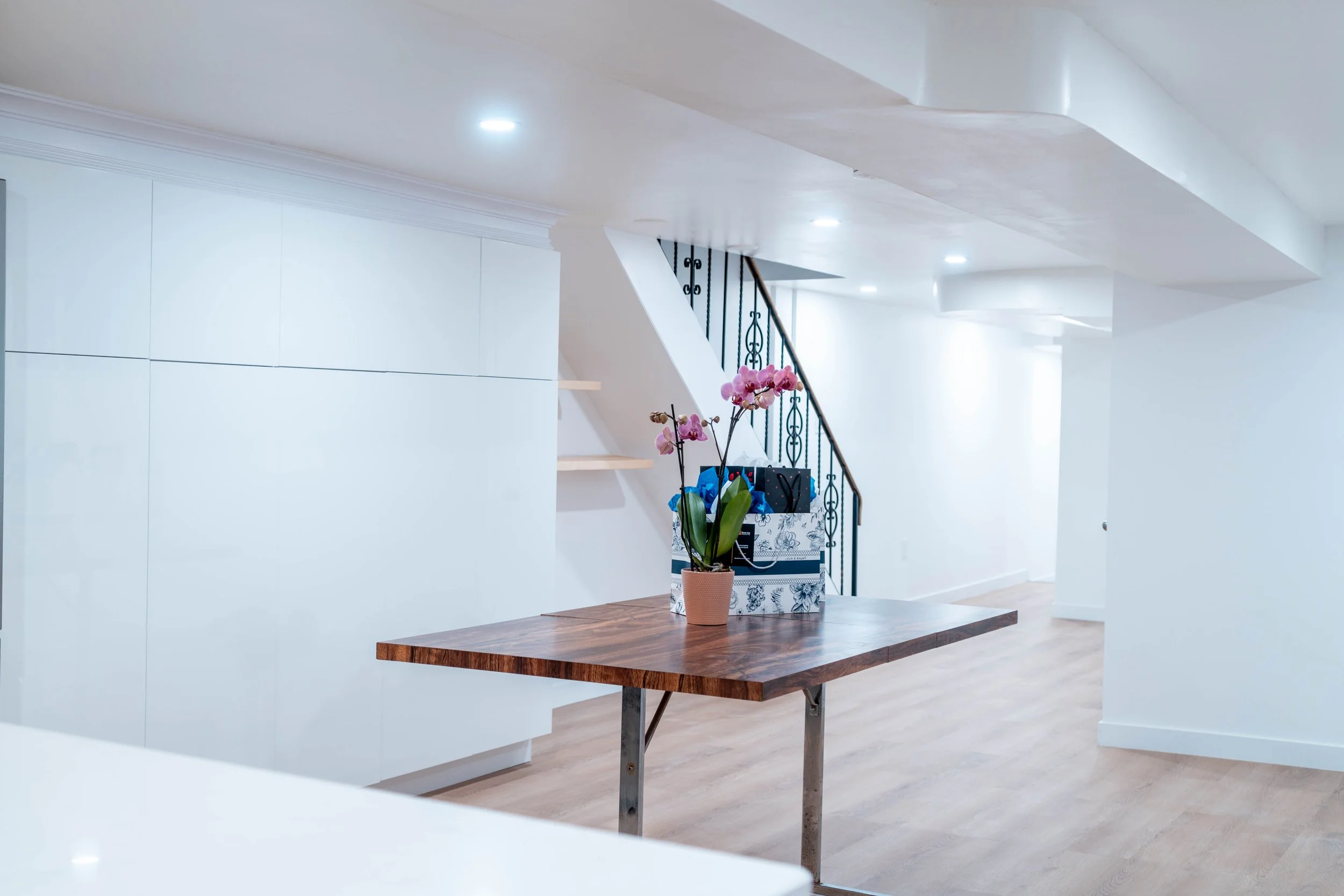 Empty living room with wooden floor, white walls, staircase with black railing, a table with a potted orchid plant, and a box with gifts.