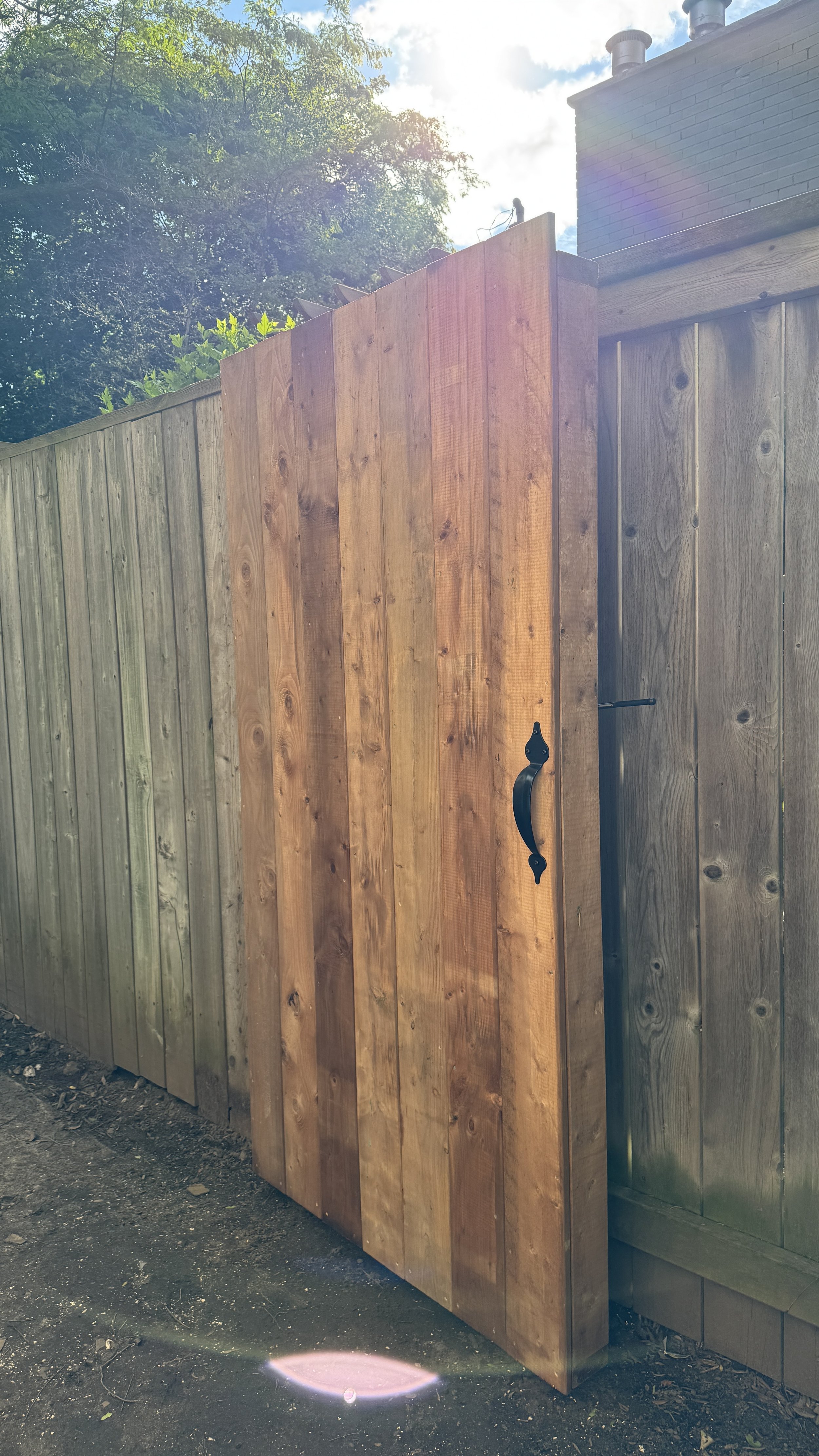 A newly installed wooden fence gate with decorative black handle and latch, adjacent to an existing wooden fence, with trees and sky in the background.