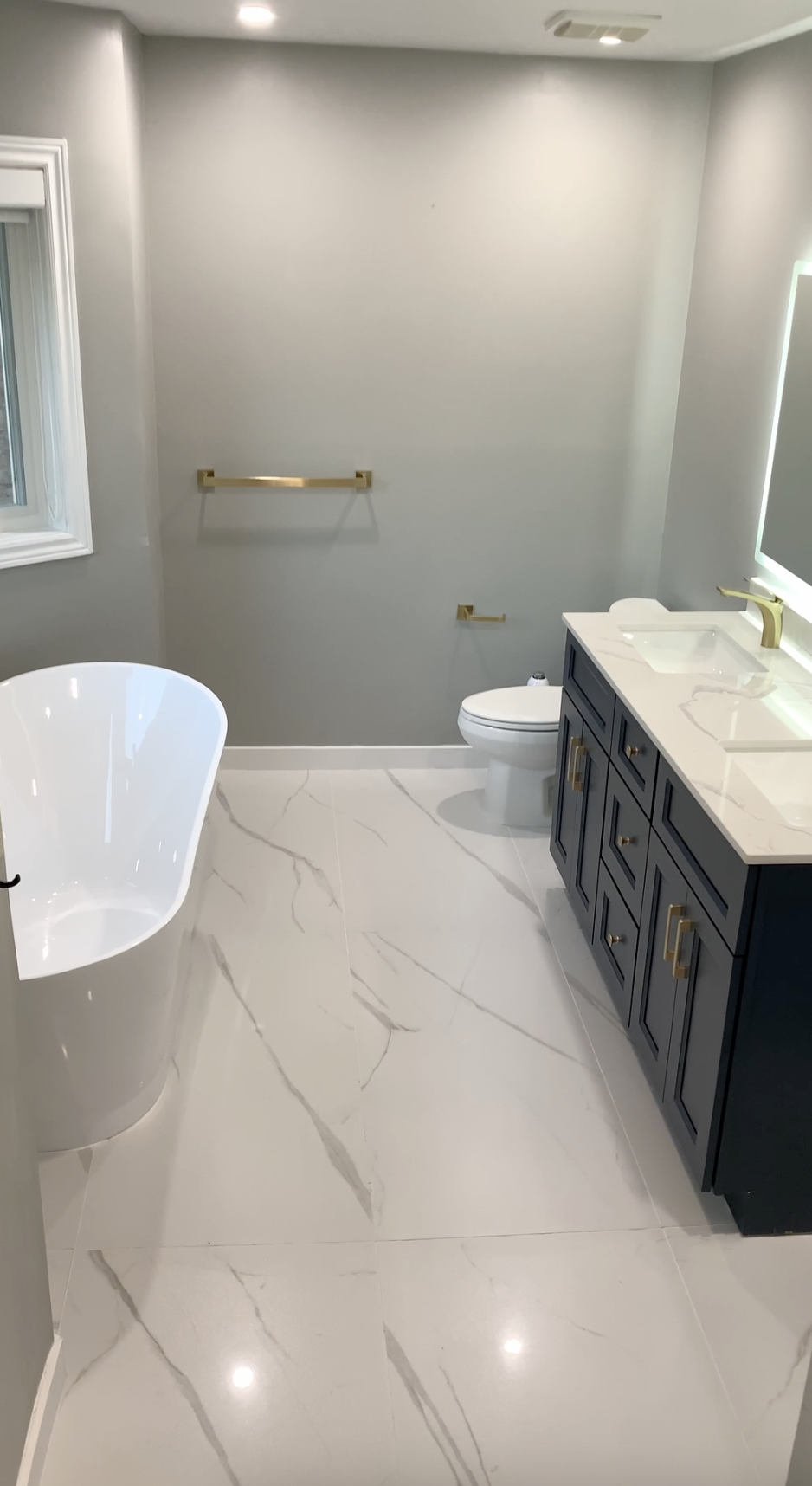 Modern bathroom with a white free-standing bathtub, a toilet, and a dark blue vanity with gold handles and fixtures. The bathroom has marble-like floor tiles and light gray walls inside a well-lit room.