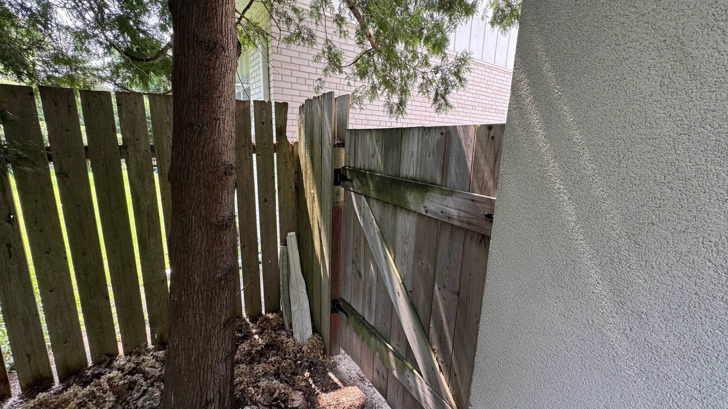 A wooden gate with black hinges and latch, partially open, next to a wooden fence and a tree, with a brick building in the background.