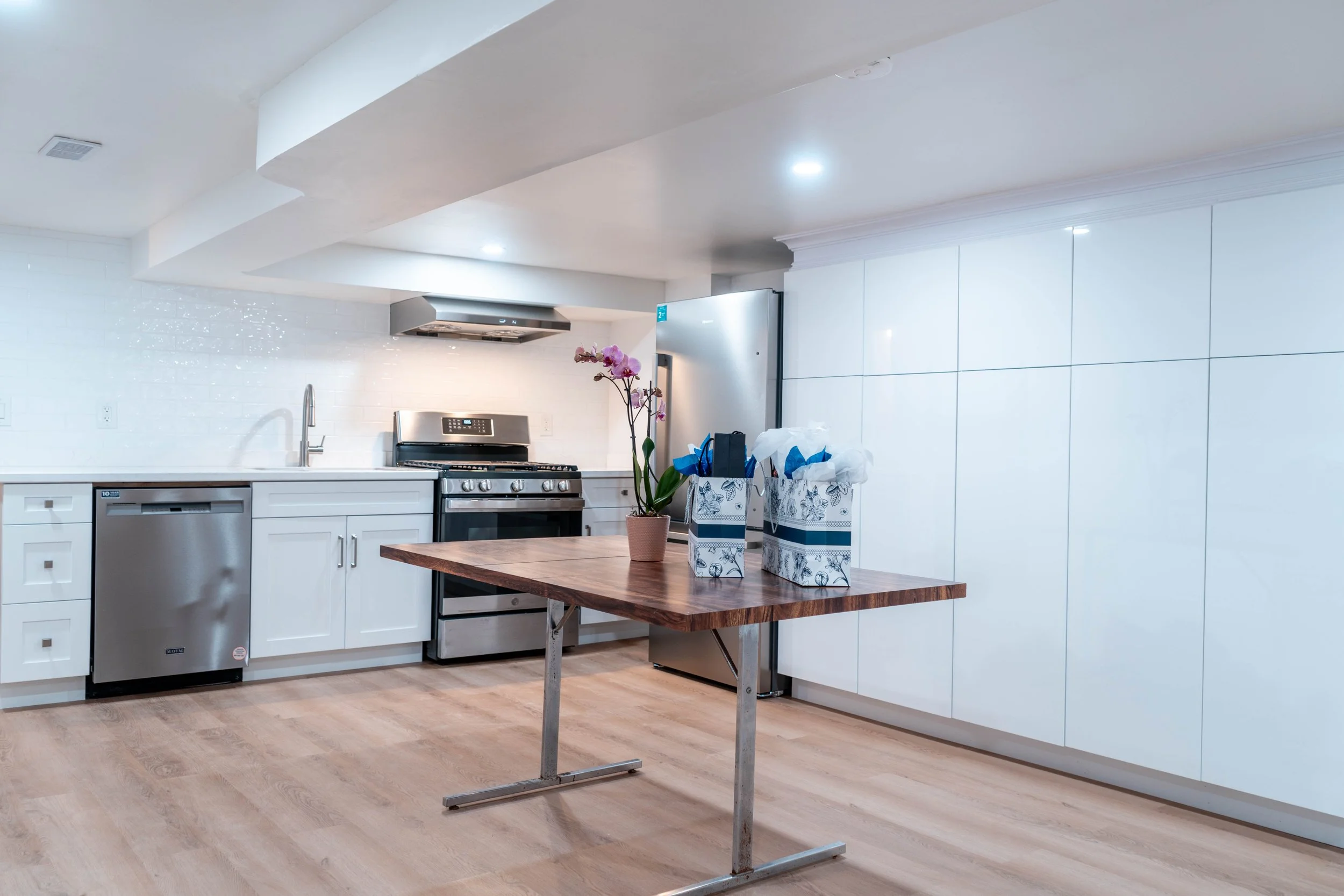 A modern kitchen with white cabinets, stainless steel appliances, a wooden table with a potted orchid and gift bags, and hardwood flooring.