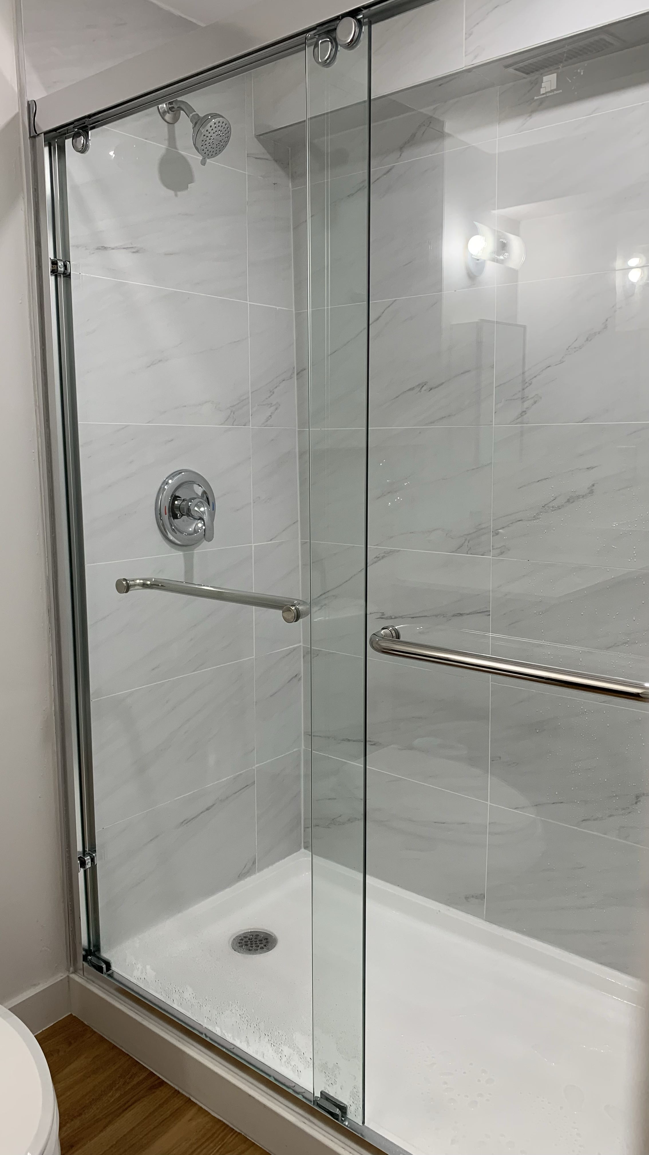 Bathroom shower with glass door, white marble tiles, and a silver handrail.