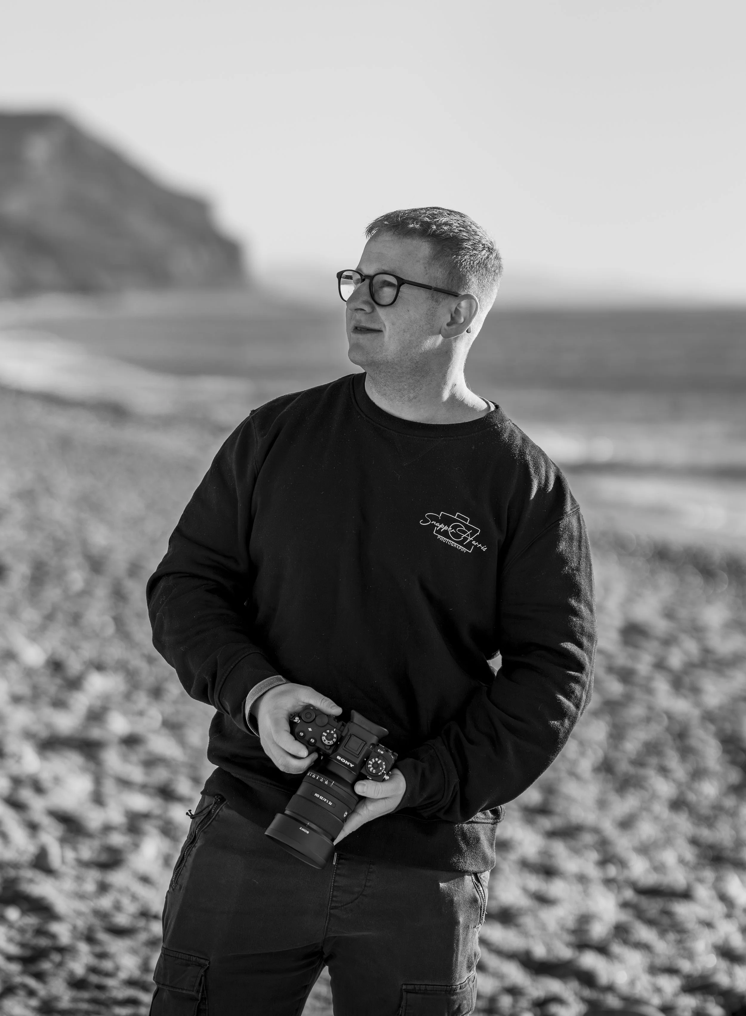 A man stands on a beach with a camera in his hand, wearing glasses and a black sweatshirt, looking off into the distance with a rocky cliff and ocean in the background.