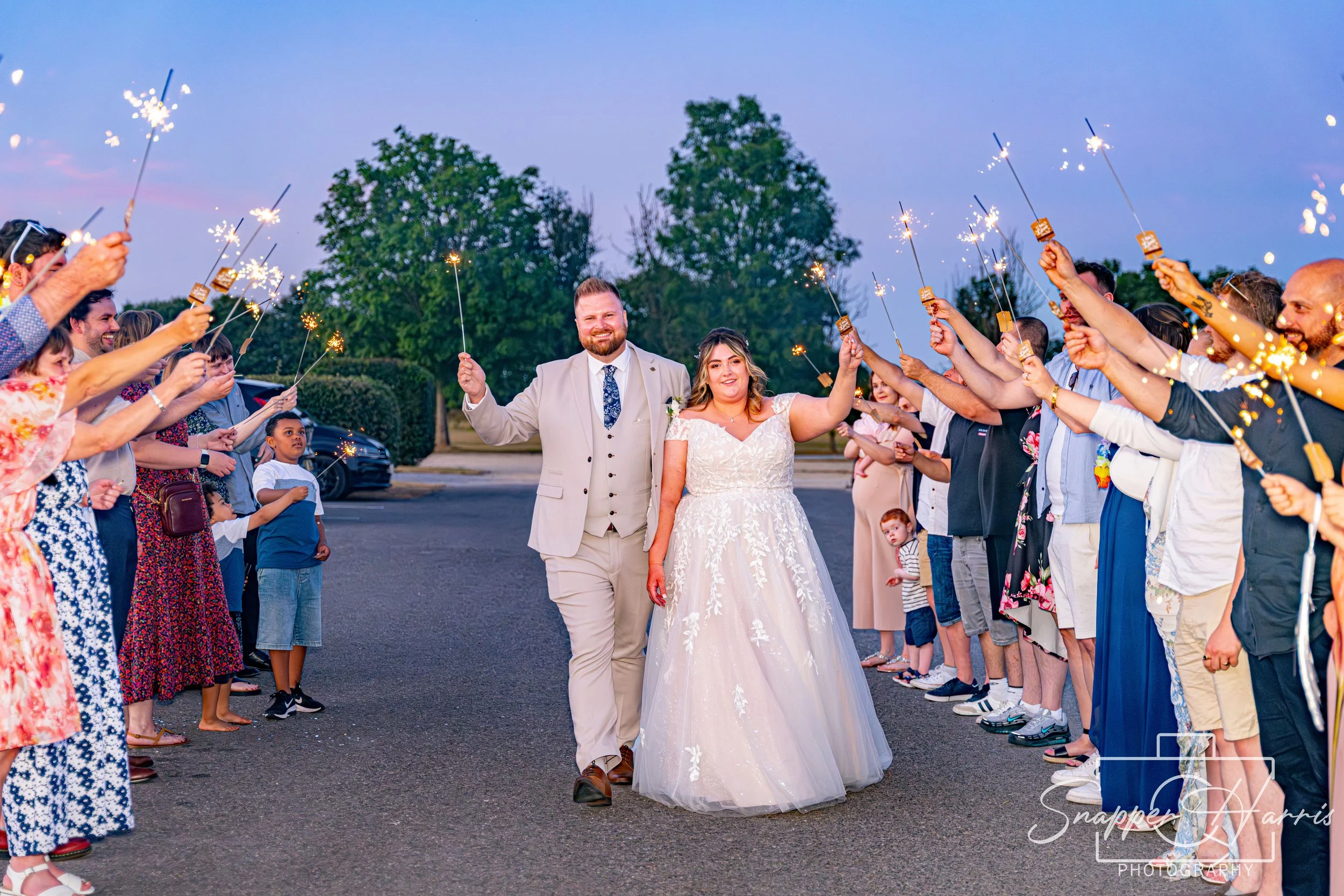 A newlywed couple walking through a sparkler exit at dusk, surrounded by friends and family on a street.