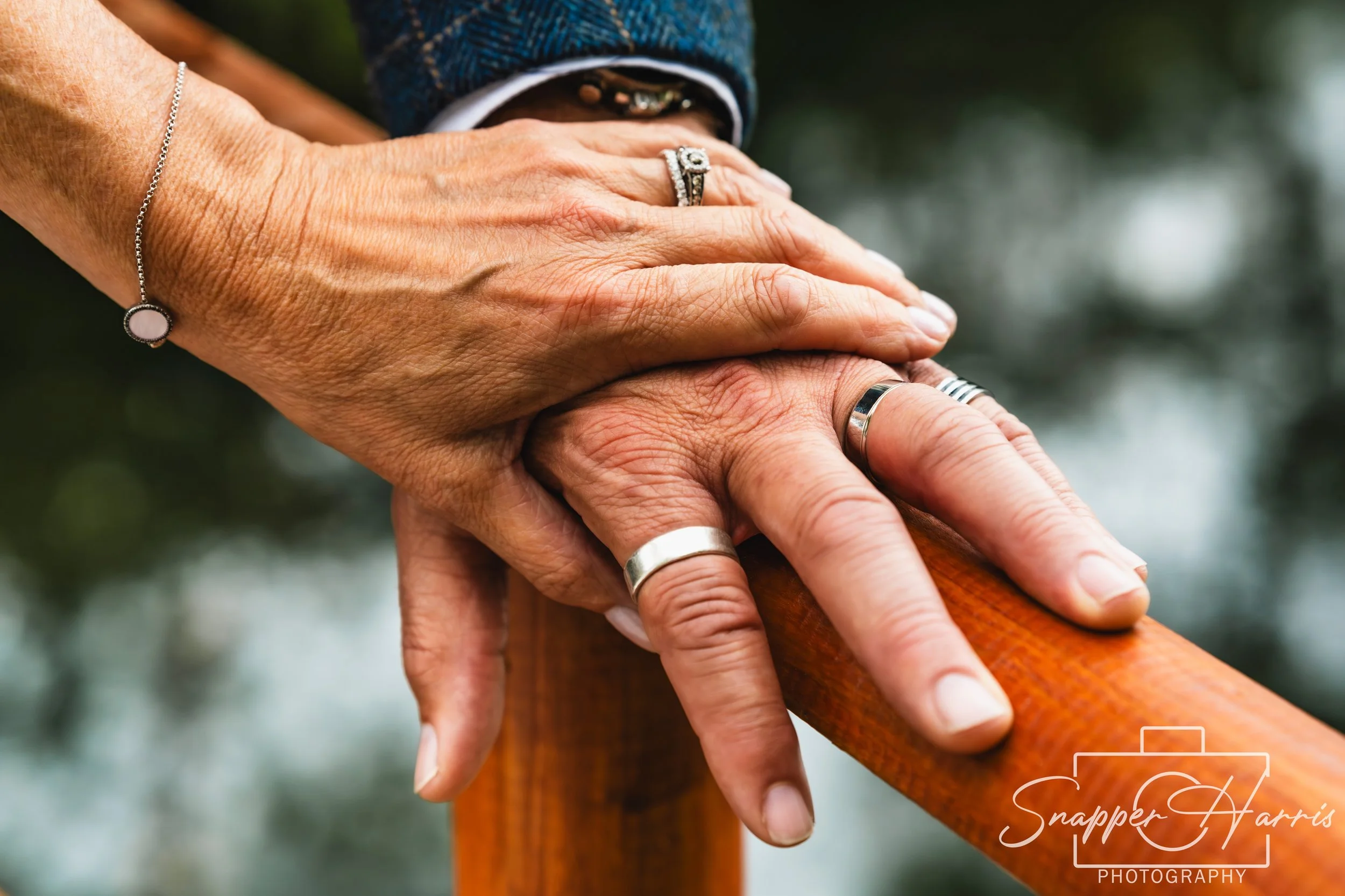 Two hands, one older and one younger, gently resting on a wooden rail. The older hand is decorated with rings, and the younger wrist is adorned with a bracelet. The background is blurred outdoors.