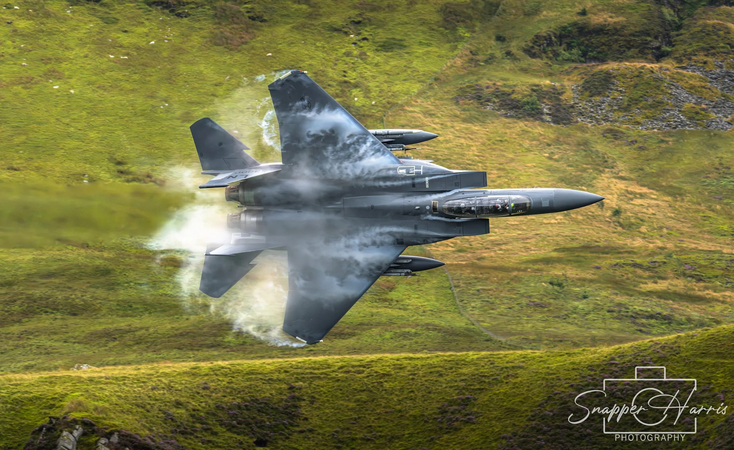 A fighter jet flying over a green grassy landscape, with vapor trails and smoke, captured from below.