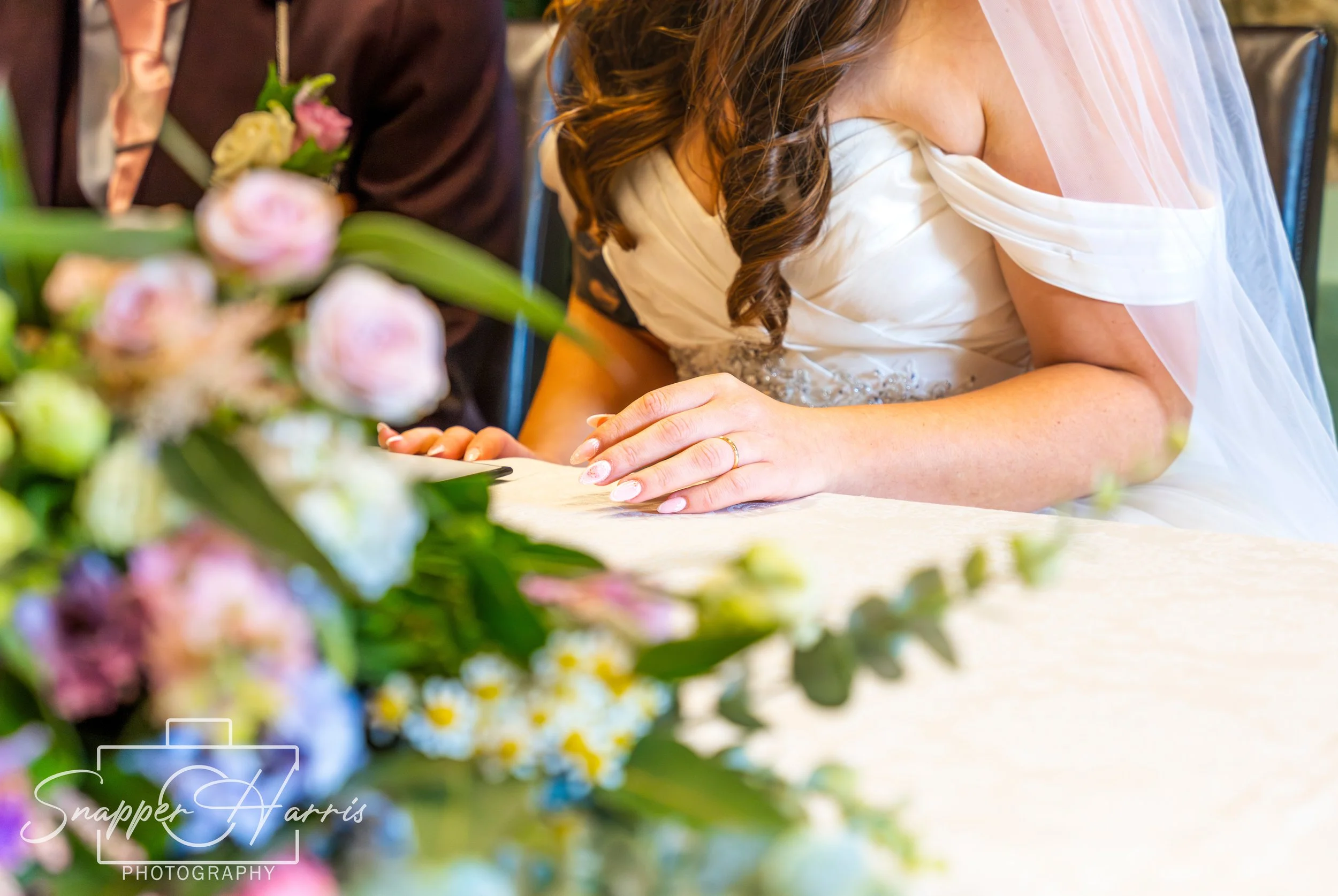 Close-up of a bride with tattooed arm and wedding ring, wearing a white wedding dress, praying during her wedding ceremony, with blurred flowers in the foreground.