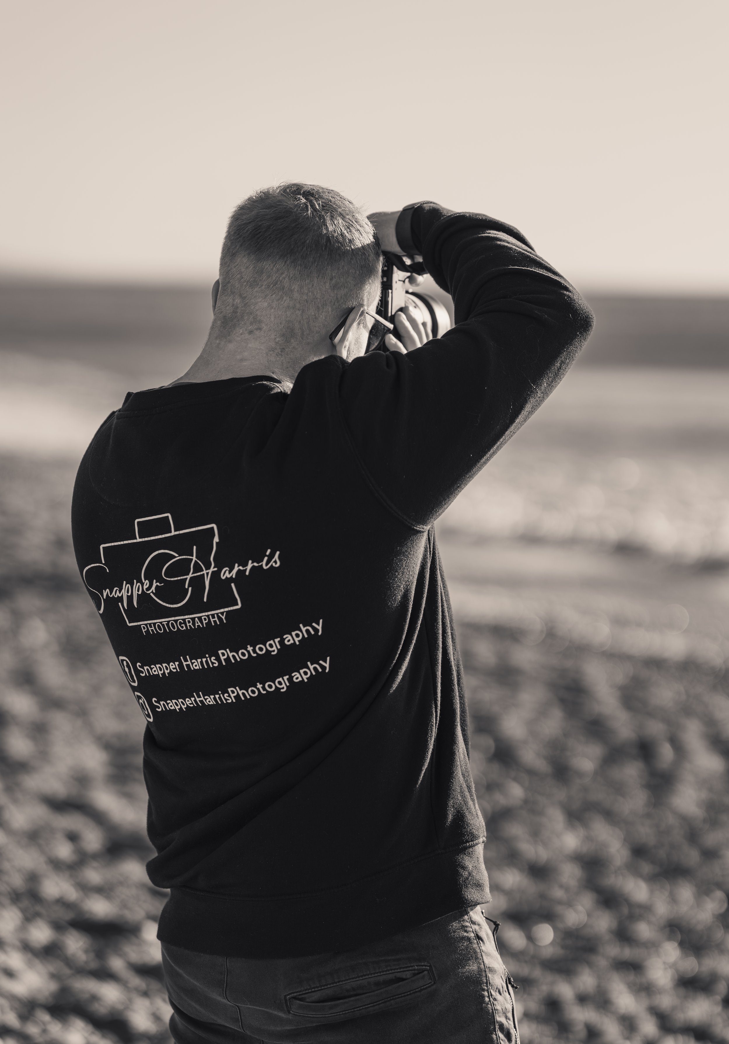 A man in a black sweatshirt taking a photo on a beach with a camera, seen from behind.