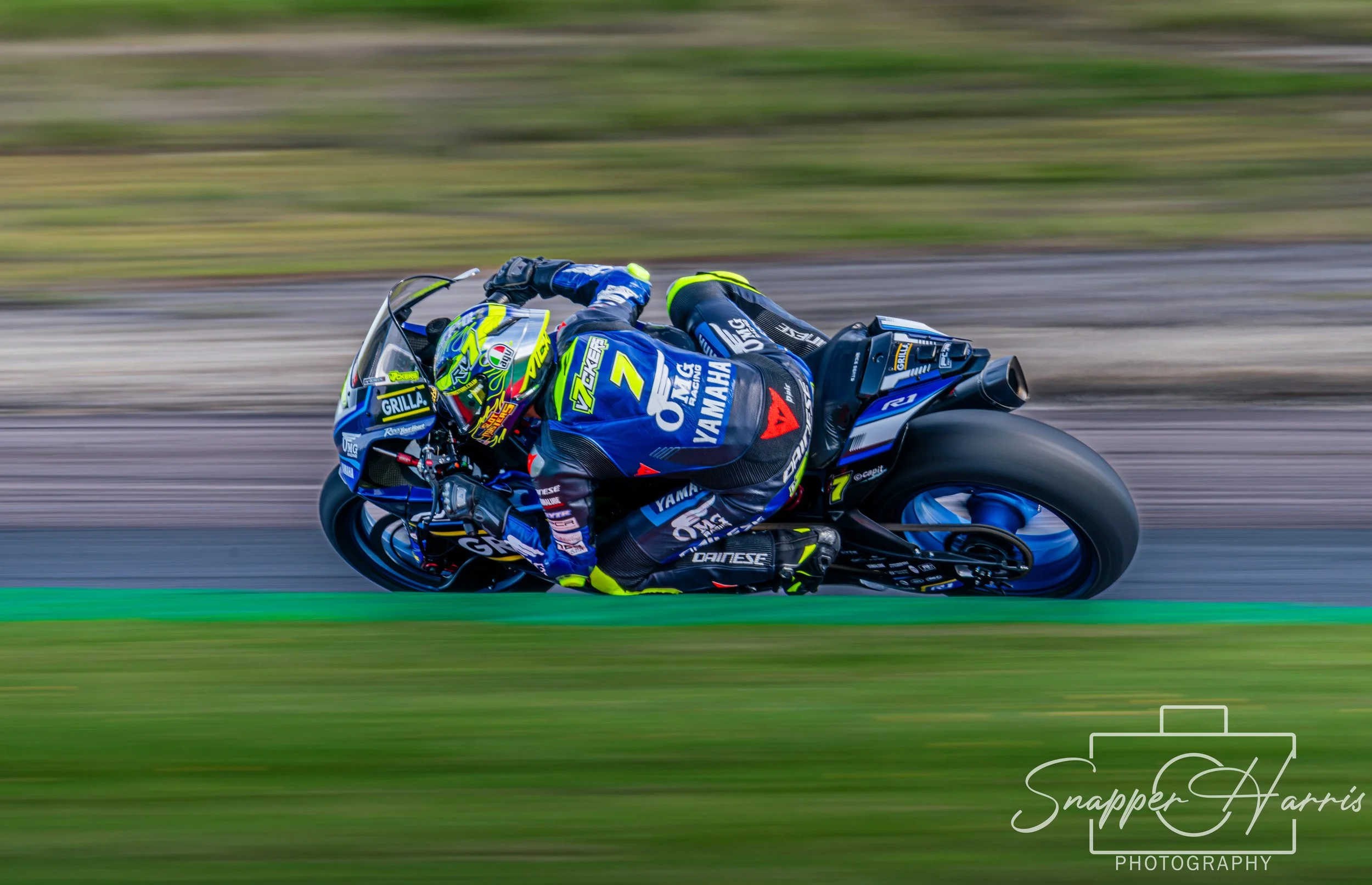 Motorcycle racer leaning into a turn on a race track, wearing a blue and black racing suit with yellow accents and a helmet, with a blurred background indicating high speed.