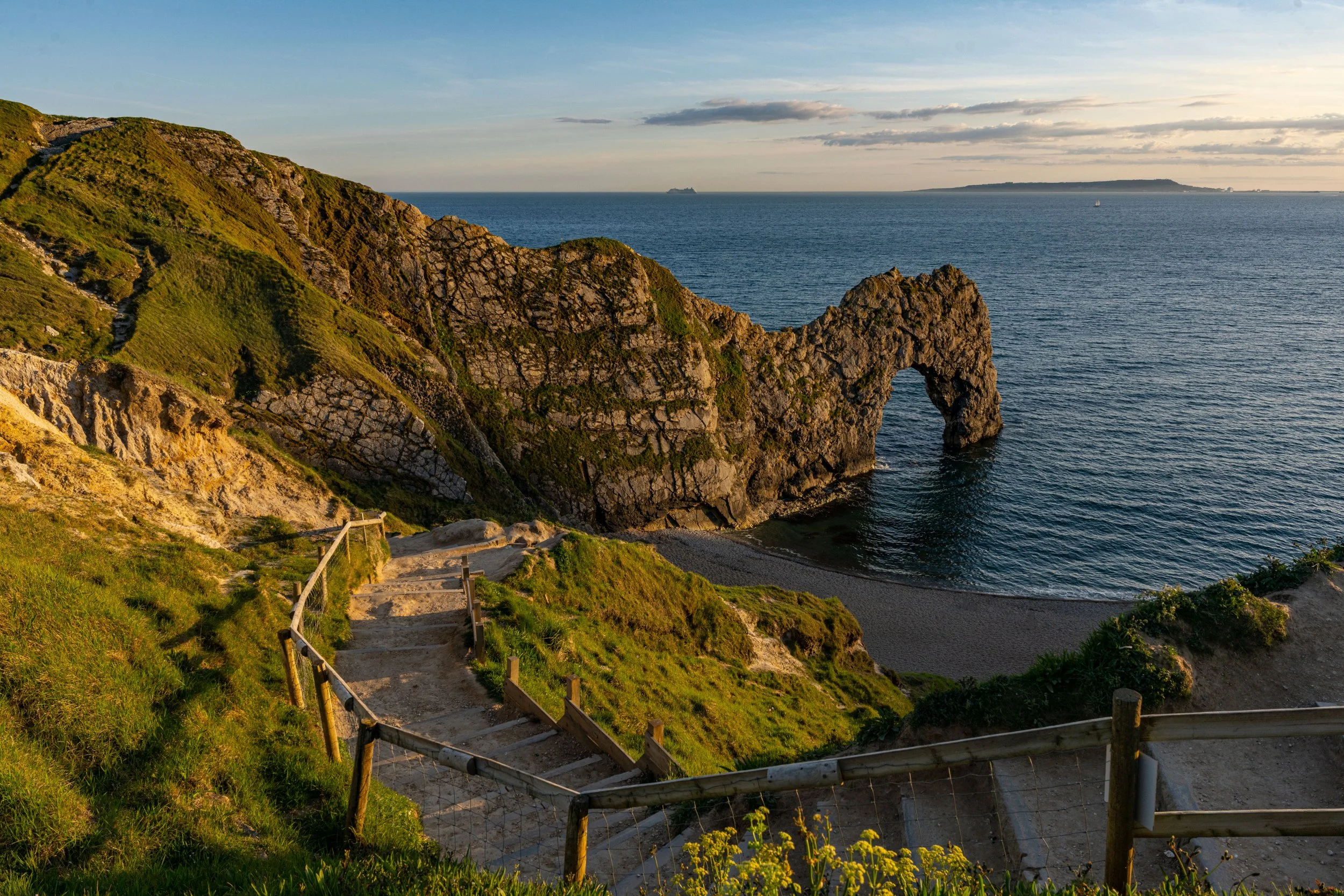 View of Durdle Door, a natural limestone arch on the Dorset coast in England, with green hillside, a winding trail, and an ocean with distant ships.