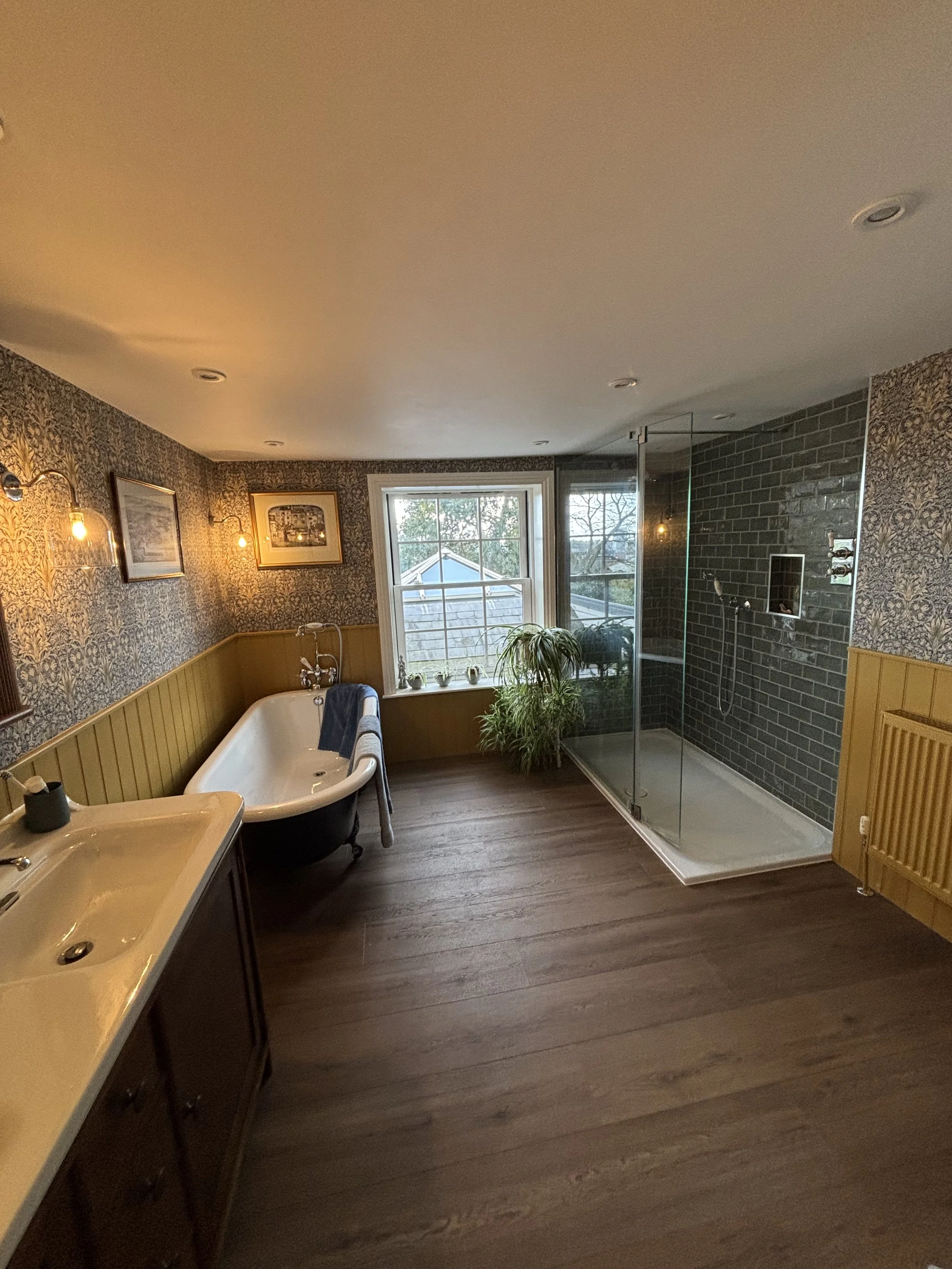 A bathroom with a tub, a window, a plant, a glass shower enclosure, and wooden flooring.