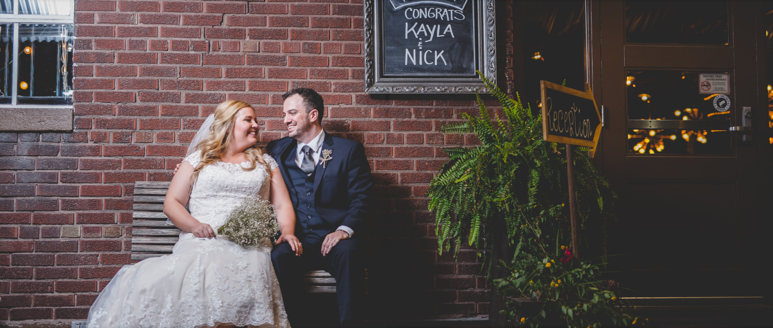 A newlywed couple sitting on a bench outside a brick building, smiling and looking at each other, with wedding attire and a bouquet of flowers.