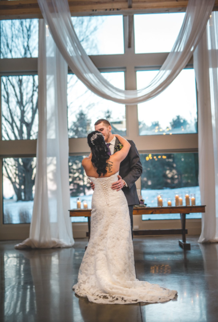 Bride and groom dancing during their wedding reception in a bright, indoor venue with large windows, flowing white curtains, and candles on a table behind them.