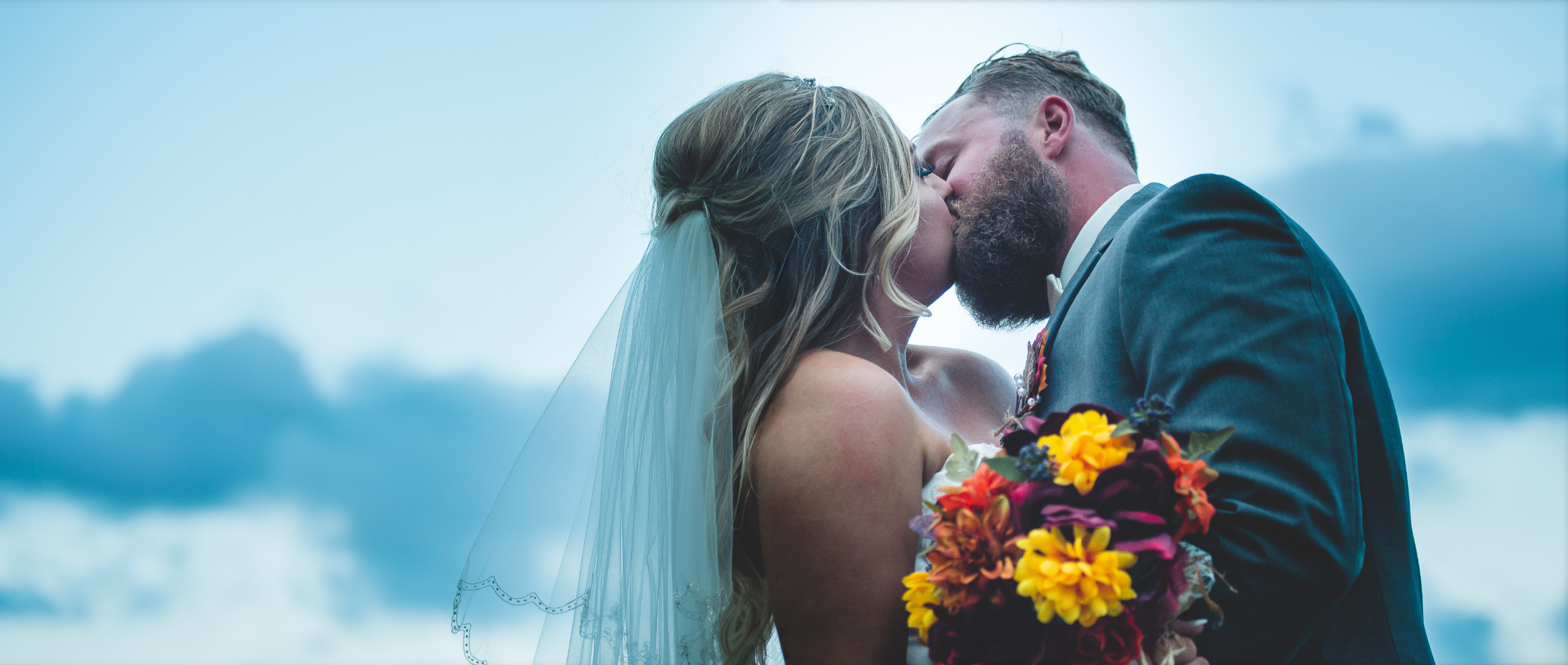 A bride and groom share a kiss outdoors on their wedding day, with the bride holding a colorful bouquet of flowers against a cloudy sky background.